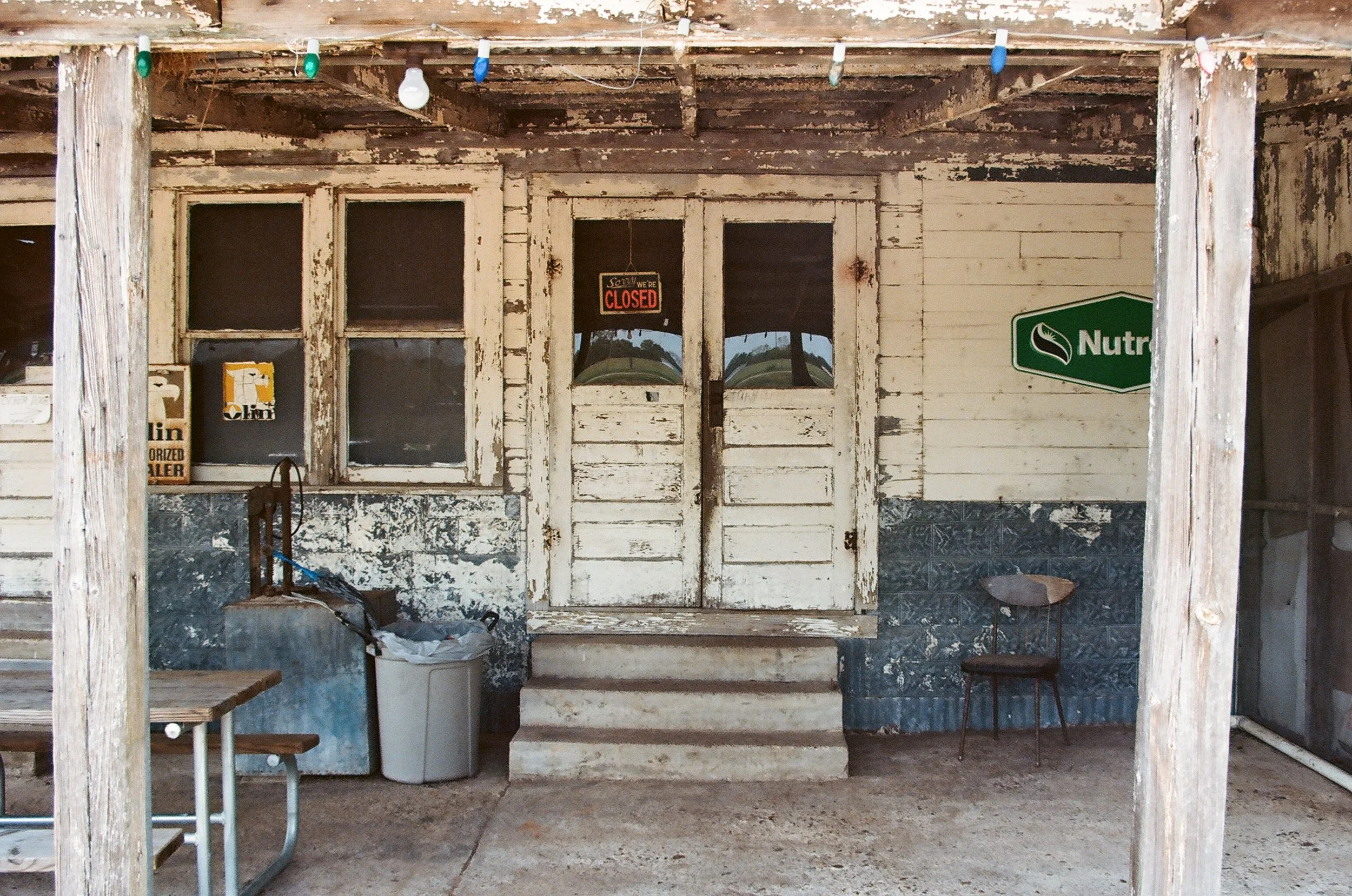 Weathered front porch with closed double doors, peeling paint, and a small set of steps, featuring a 'Sorry we're closed' sign, an old chair, a trash bin, and a partly visible sign reading 'Nutri' on the right.