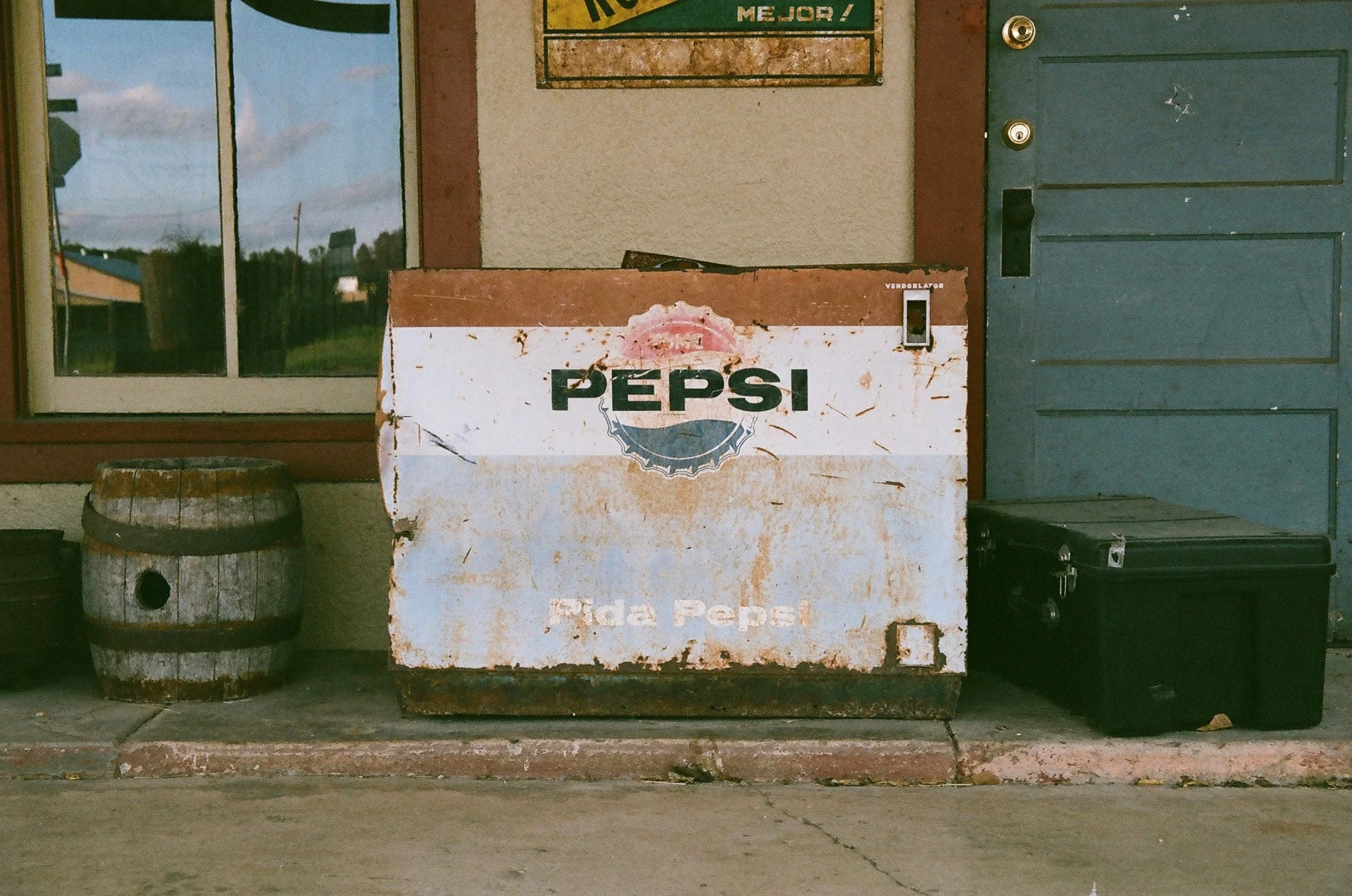 Old, weathered Pepsi cooler with peeling paint and rust, placed outside a building next to a wooden barrel and a black toolbox. A window with reflected sky and a door are in the background.