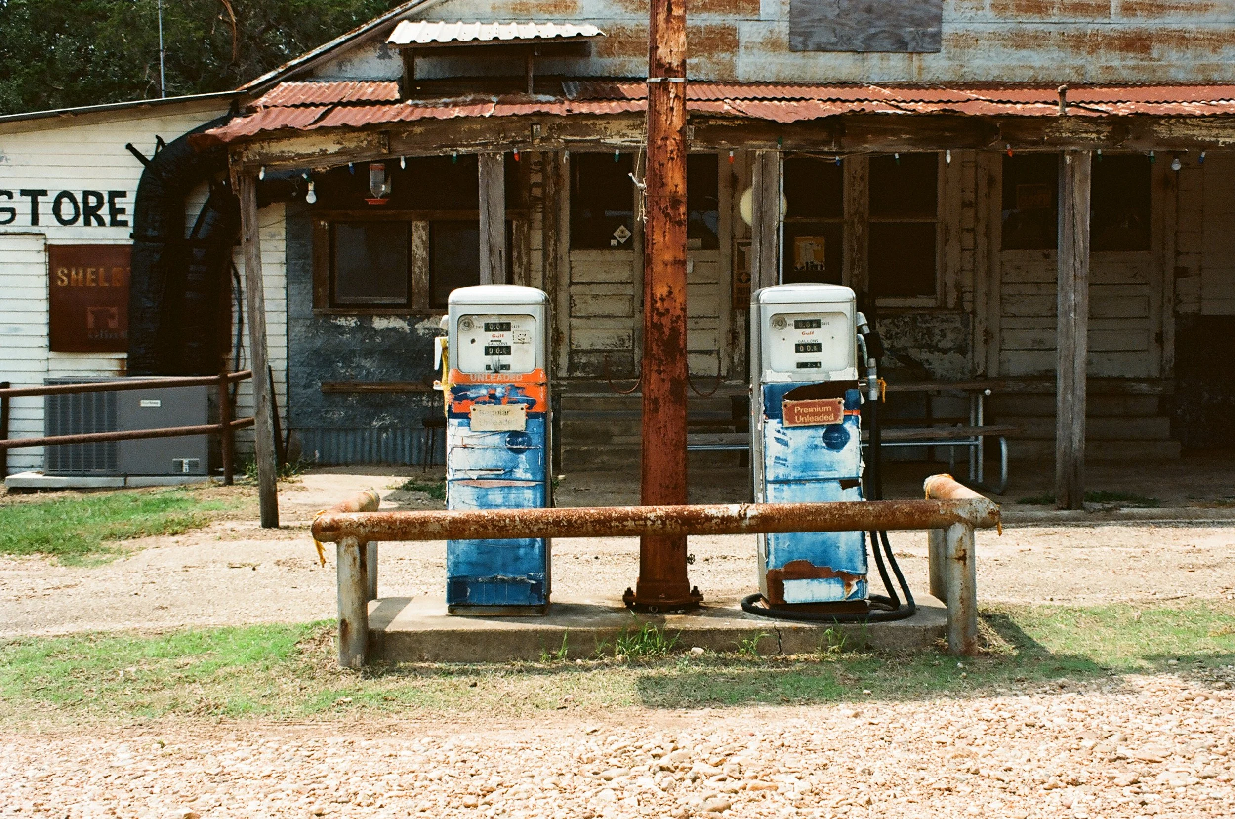 Old, rusted gas pumps in front of a weathered, wooden building with a corrugated metal roof and string lights, on a dirt and grass patch.