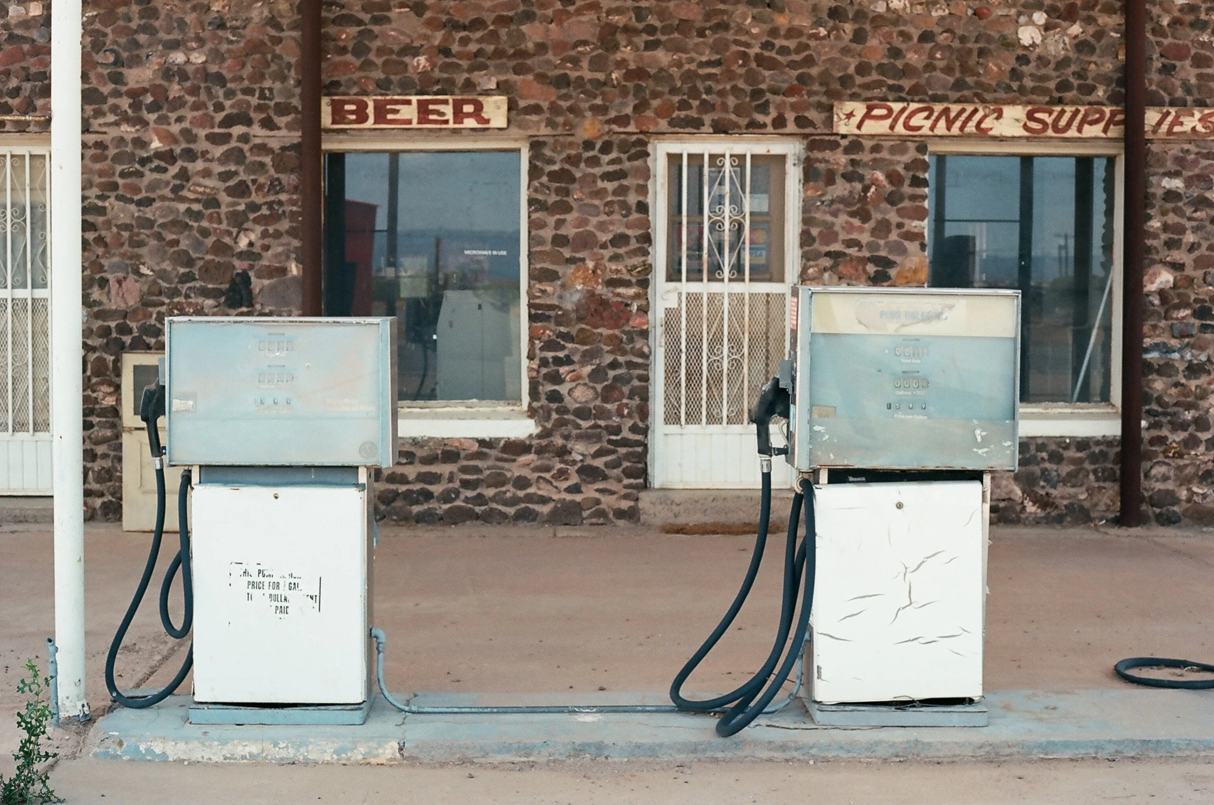 Two old fuel pumps in front of a stone building with signs for beer and picnic supplies.