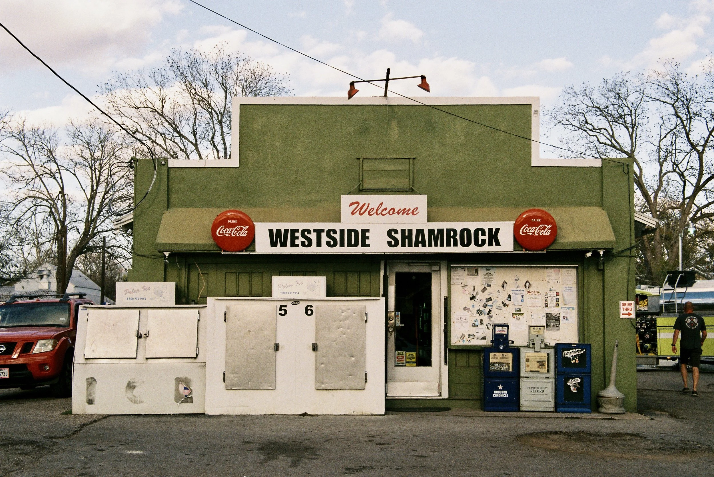 Green building with a sign that reads 'Welcome Westside Shamrock.' Two red Coca-Cola logos are on either side of the sign. The building has a small entrance door, and various objects and notices on the front, with a newspaper box and a fire truck in 