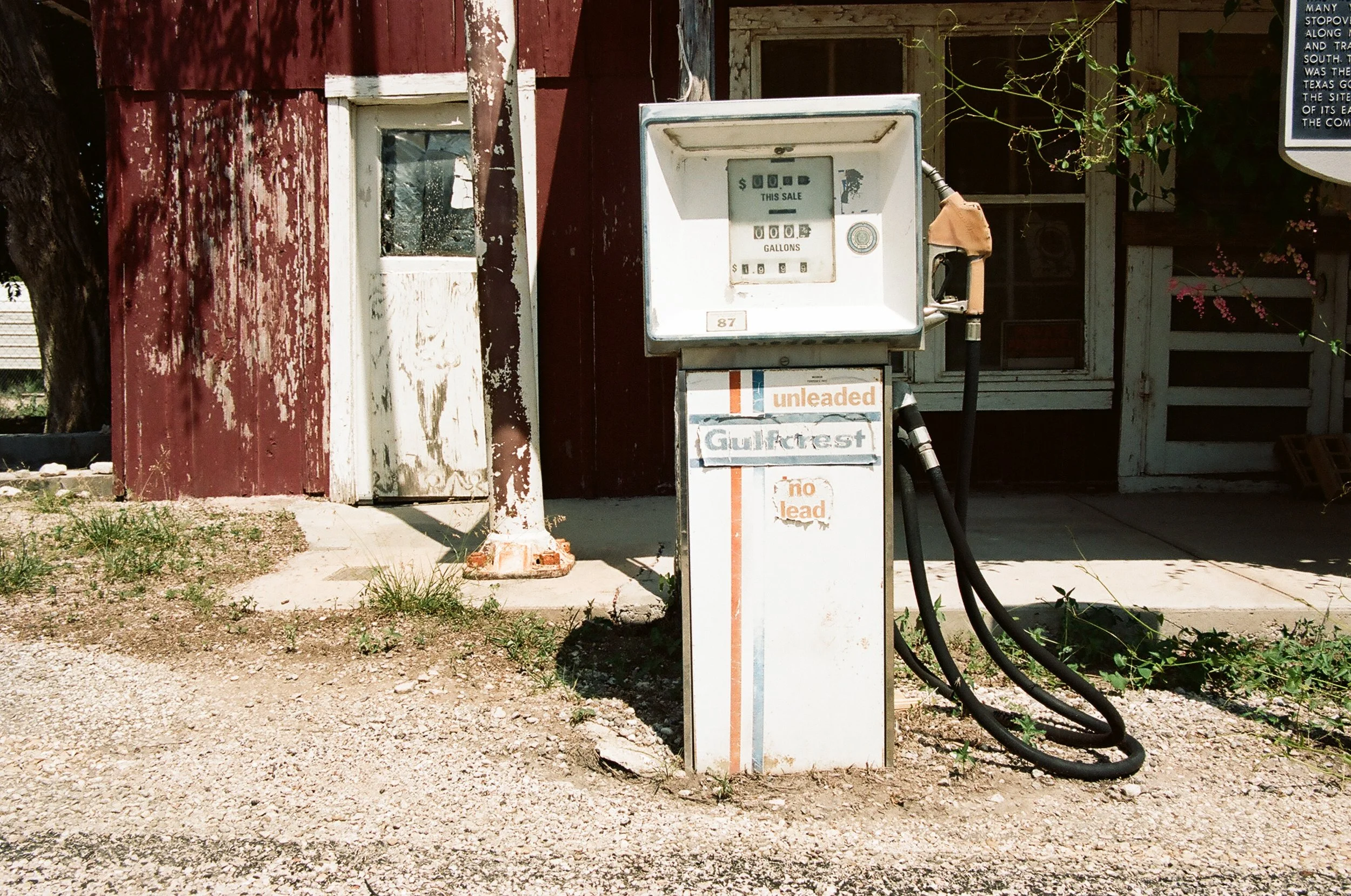 Old, weathered gas pump with peeling paint and black hoses in front of a rustic, red, weathered building with a small window and overgrown plants.
