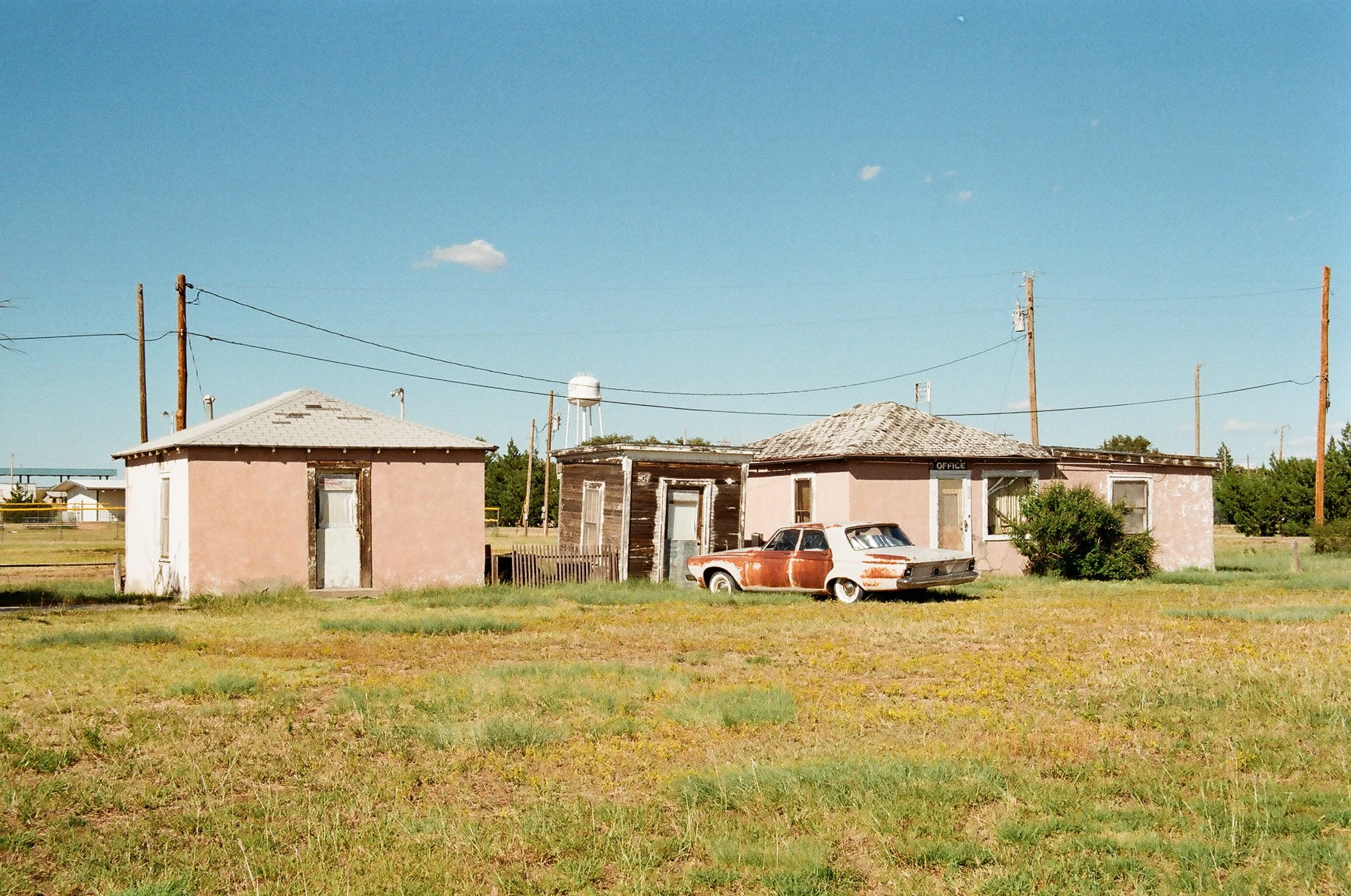 Old, weathered buildings with peeling paint and a rusty car parked in front, set in a grassy field under a clear blue sky.