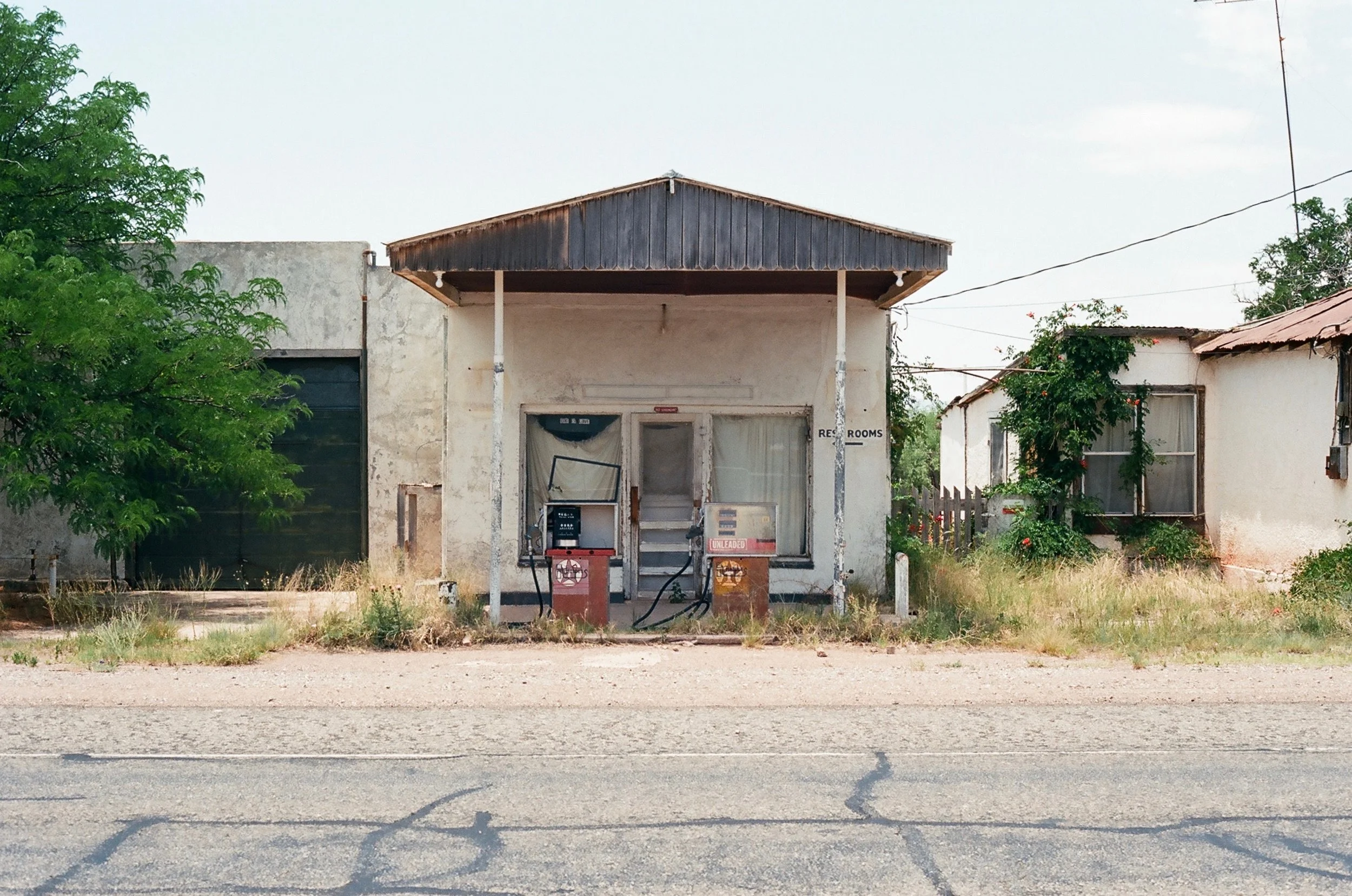 Old, abandoned gas station with two empty pumps, surrounded by overgrown grass, and small, run-down houses with peeling paint on either side.
