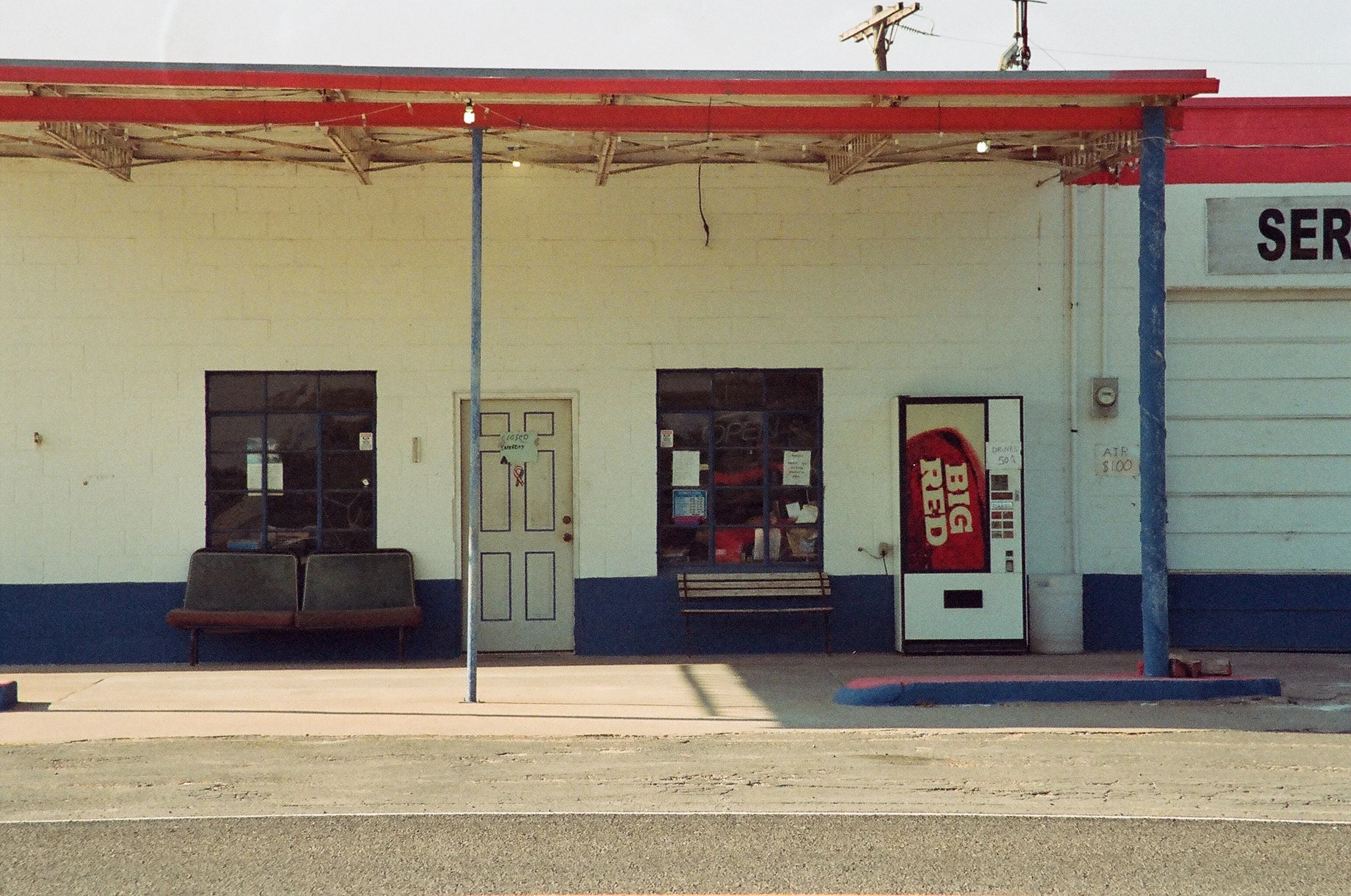 A small roadside service station with a bench, vending machine, and a door, under a red and blue awning.