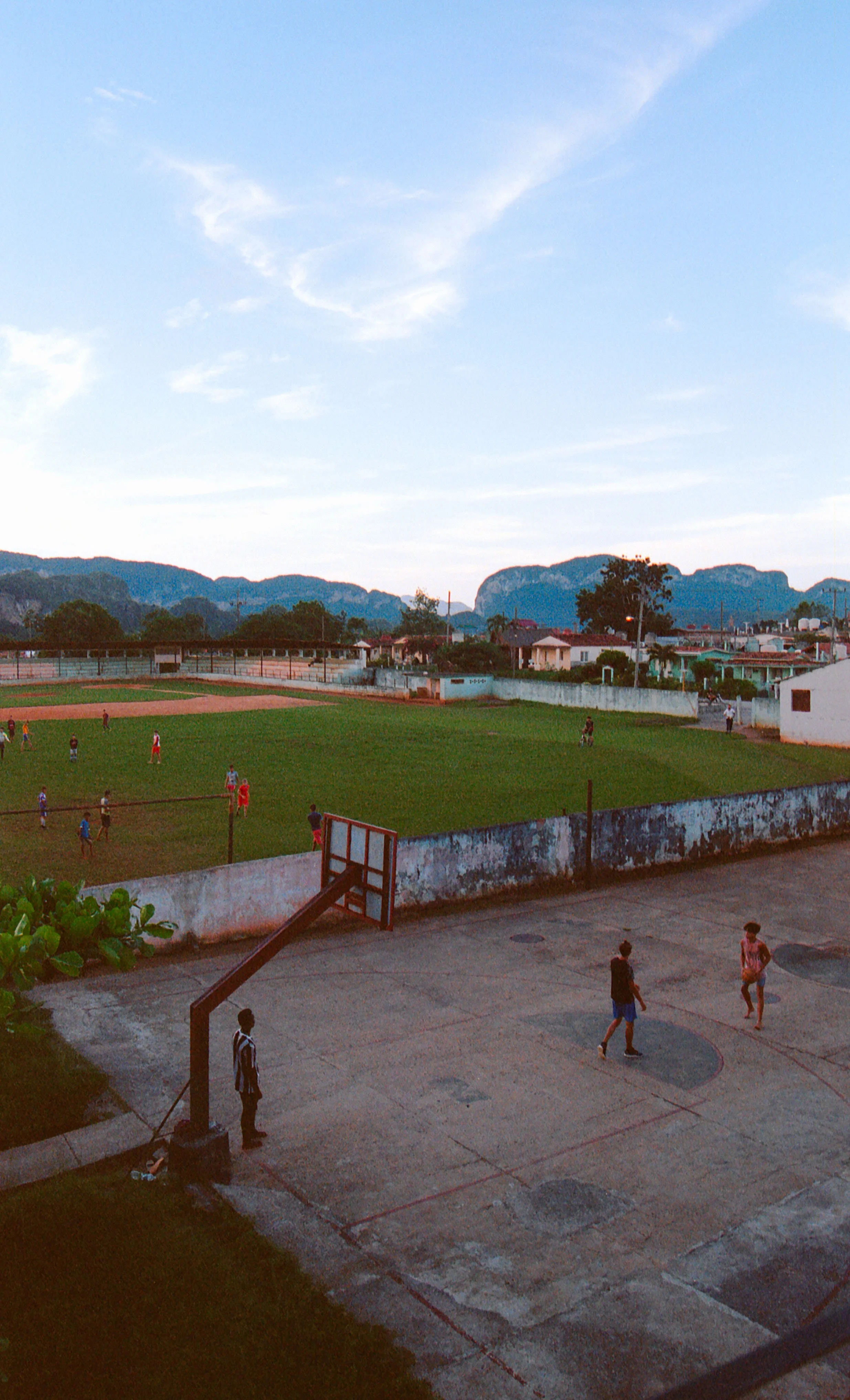 A small outdoor basketball court with players, adjacent to a baseball field where children are playing, with residential houses and mountains in the background under a partly cloudy sky.