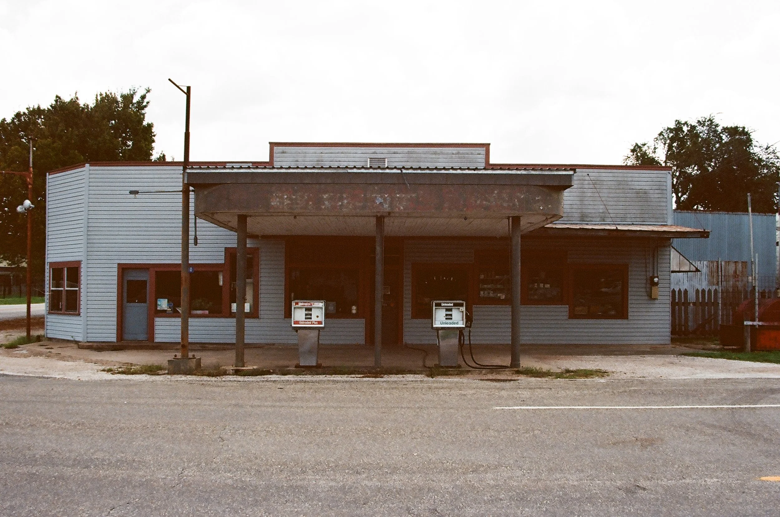 An old, weathered gas station with two fuel pumps in front, modern-sized windows, and a canopy with rusted edges, located on a street with trees in the background.