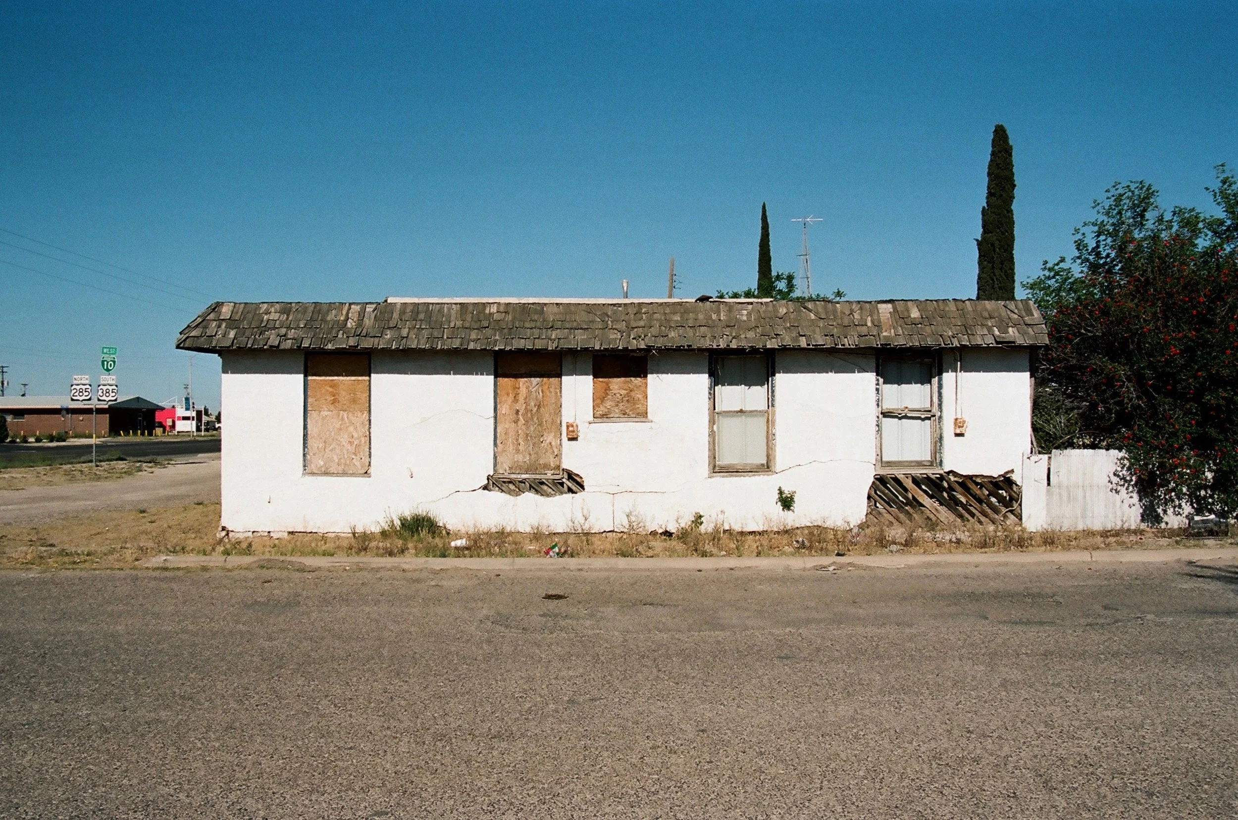 An old, abandoned house with peeling white paint, boarded-up windows, and a sagging shingle roof, situated on a deserted street under a clear blue sky.