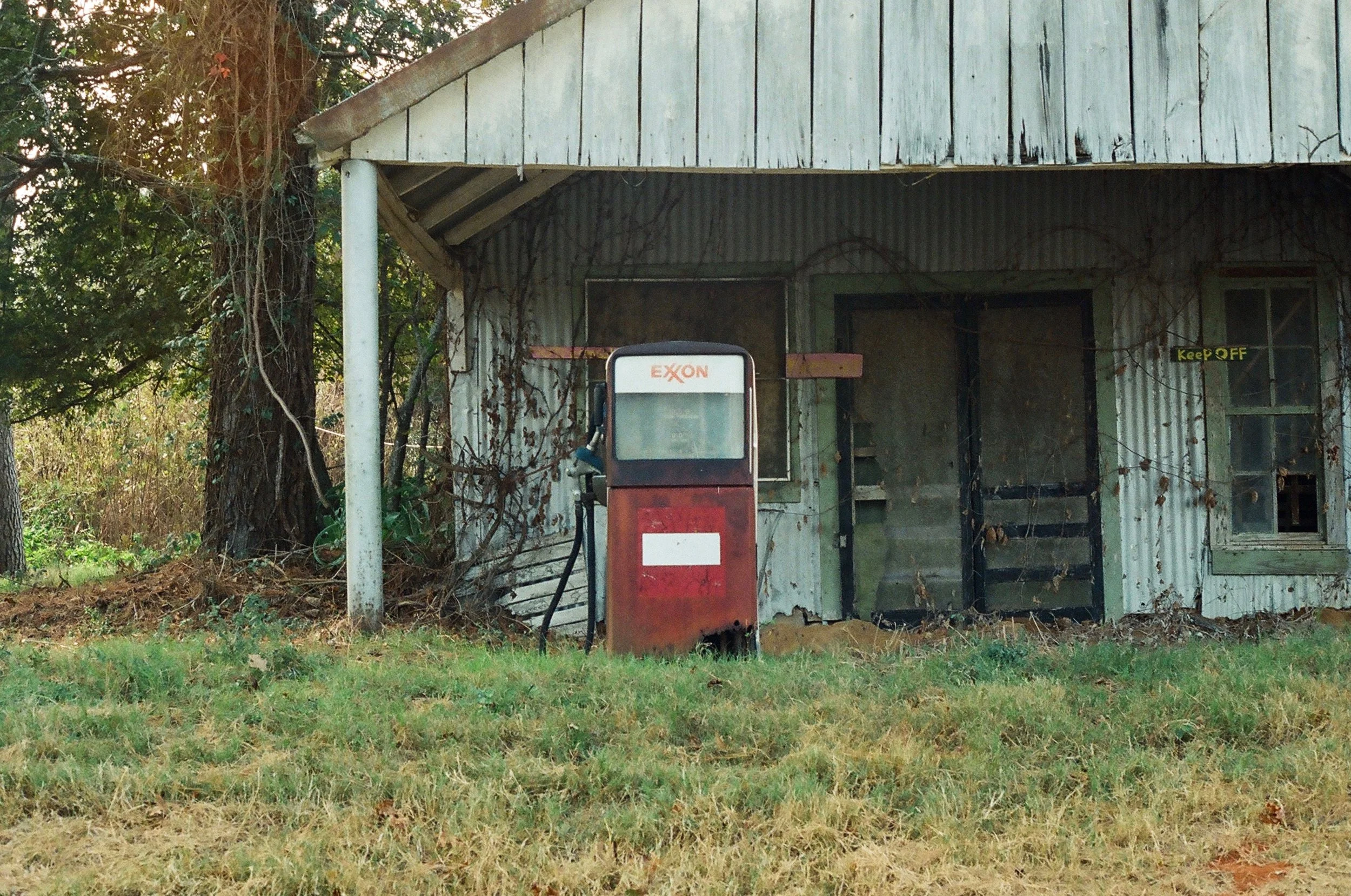 Old, rusty Exxon gas pump outside a dilapidated, weathered building with peeling paint, surrounded by grass, trees, and overgrown vines.