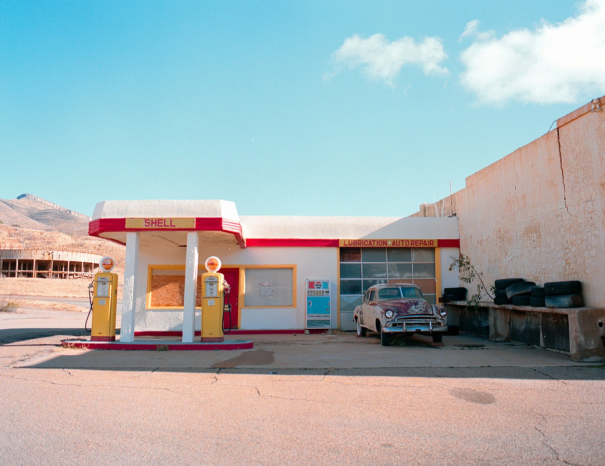Old Shell gas station with two vintage pumps, an abandoned car, and tires stacked on a concrete surface under a blue sky.