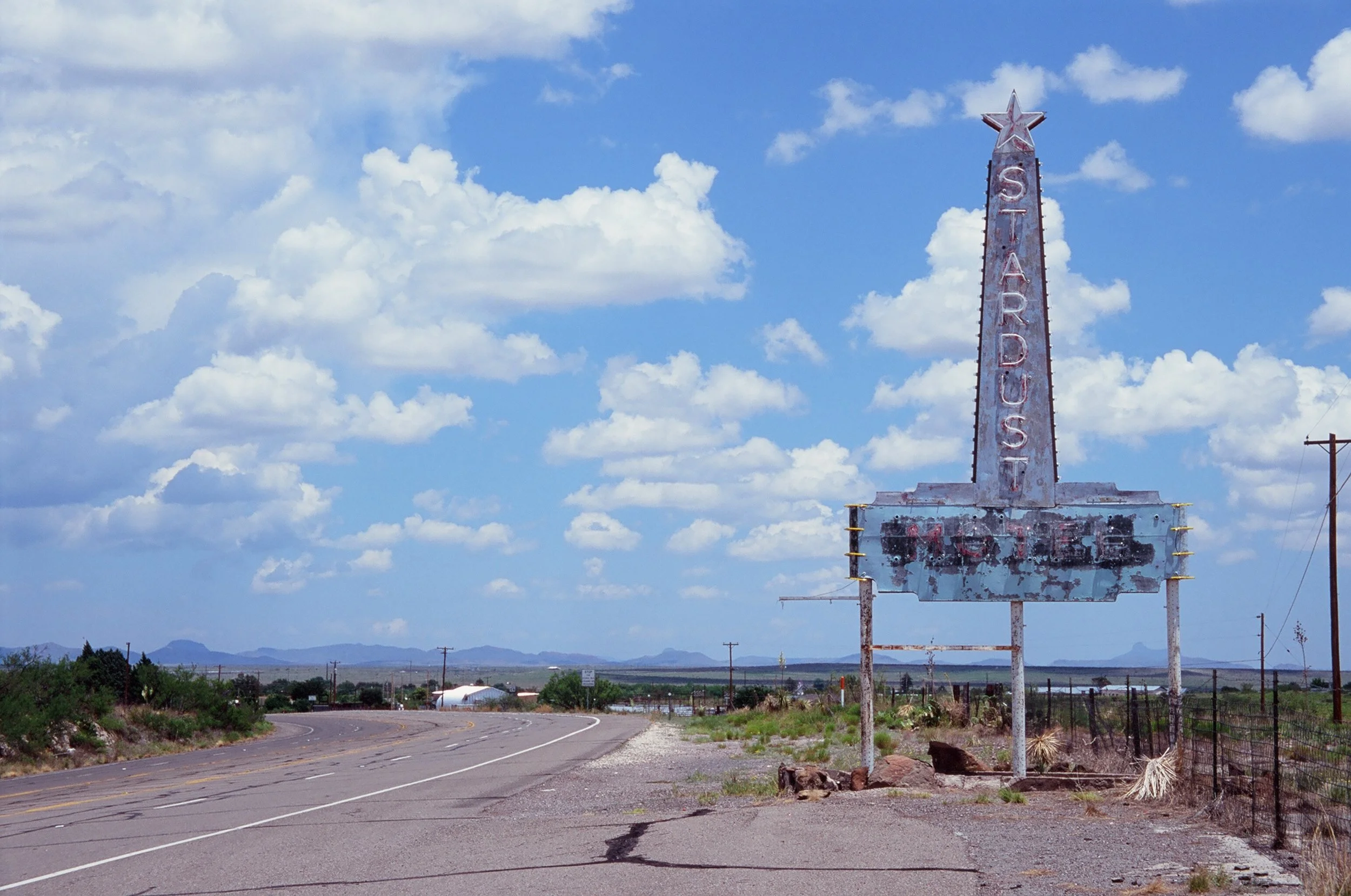 Old, weathered motel sign in a rural area with clear blue sky and scattered clouds, distant mountains, and a winding road.