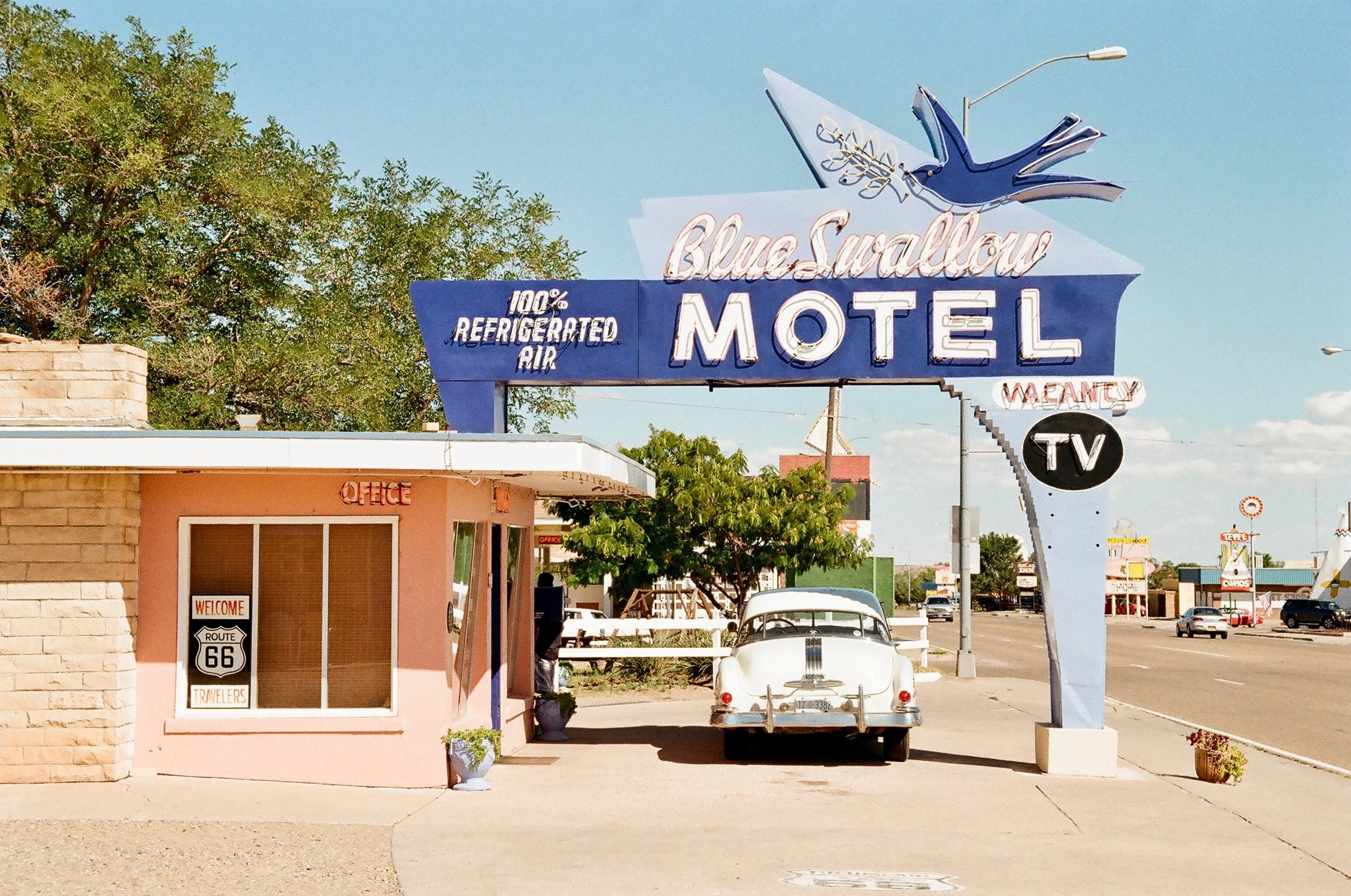 The entrance to a vintage-style motel called Blue Swallow Motel with a large neon sign. The sign features a blue swallow bird, the words "Blue Swallow," and a black circle with "TV" inside. There is a small pink office building with a window displayi