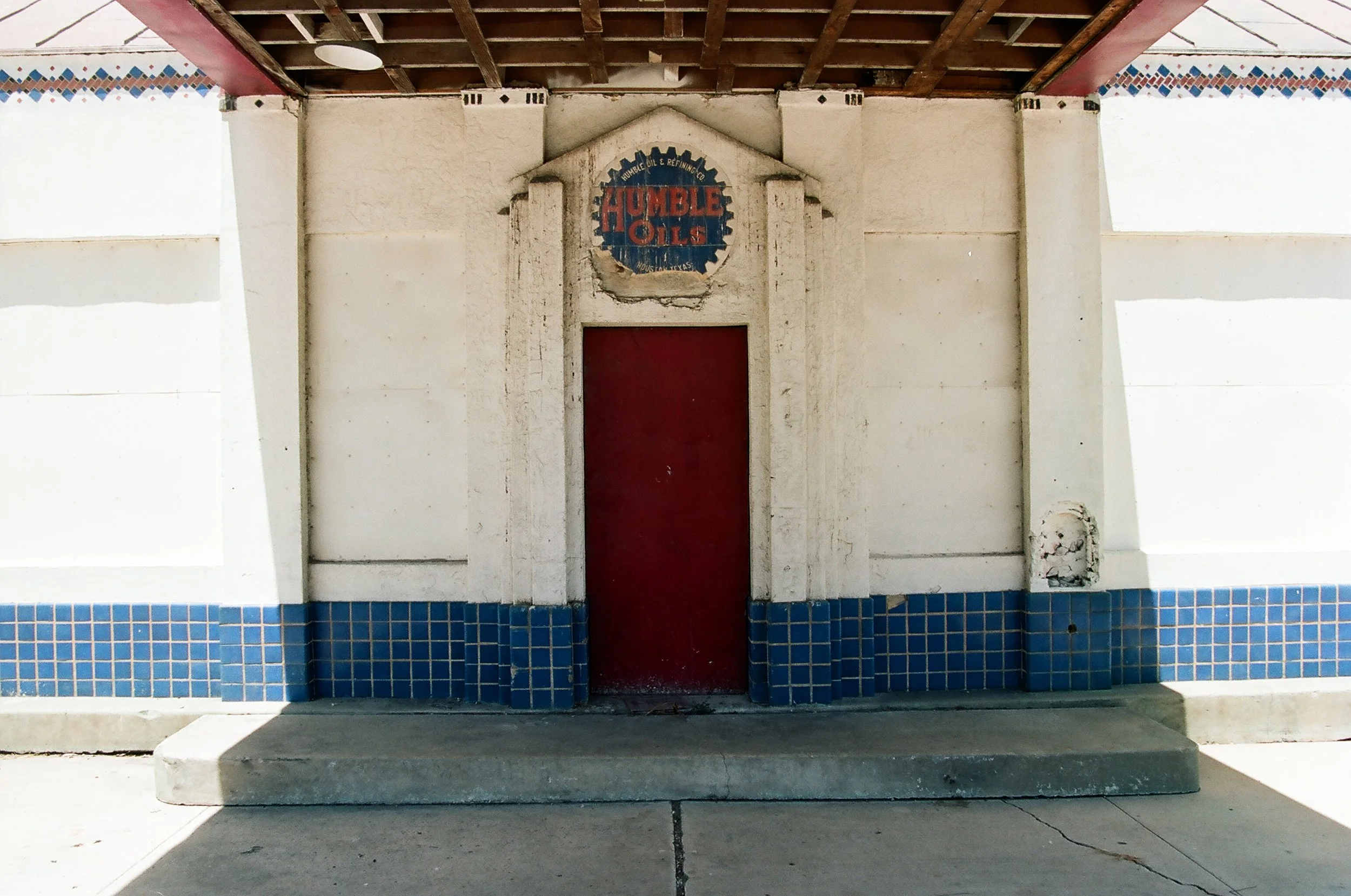 Old building with a red door, white walls, blue tile bottom trim, and a faded sign reading 'Humble Oils' above the door.