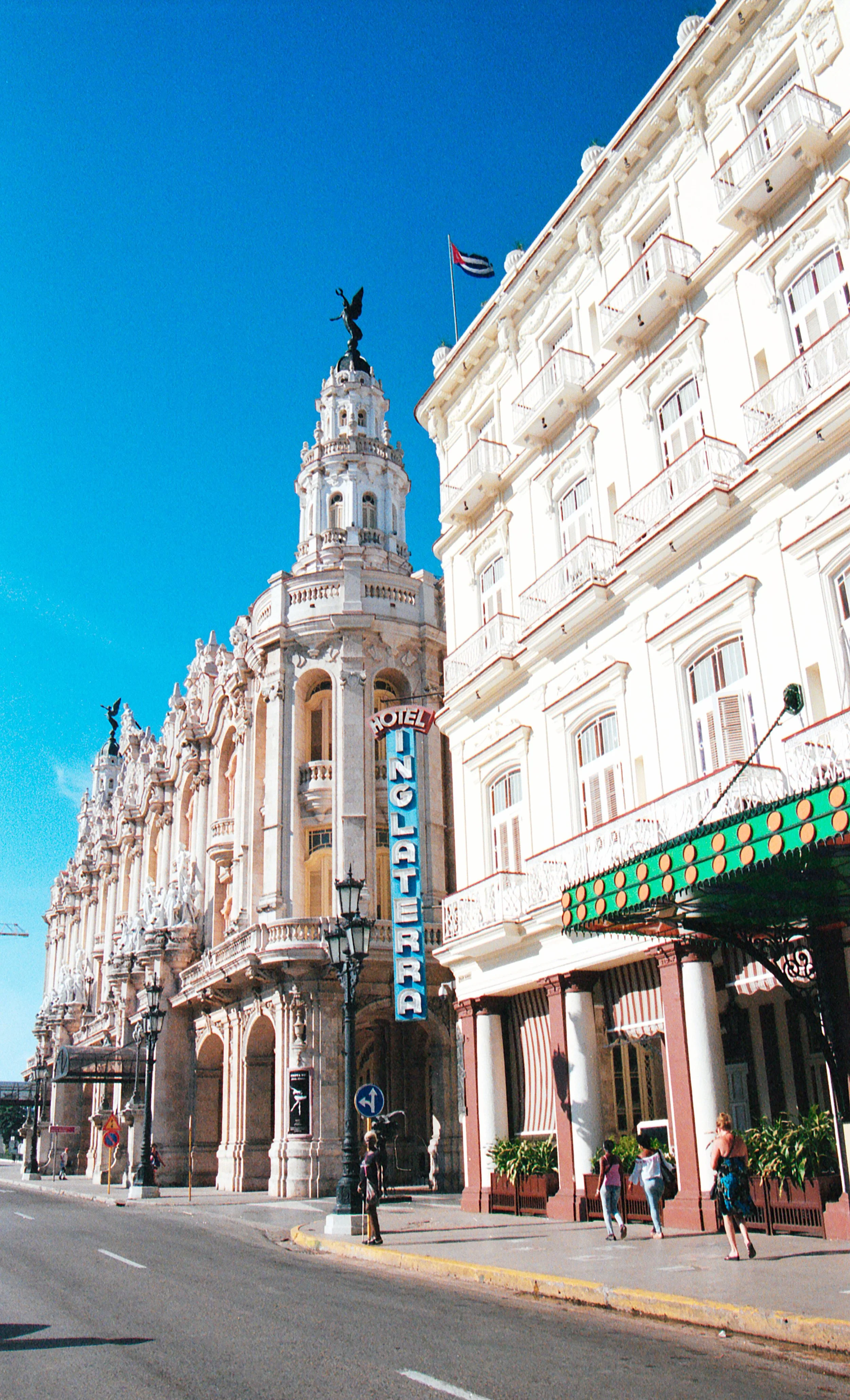Street view of a historic building with ornate architecture, a hotel sign, and people walking on the sidewalk under a bright blue sky.