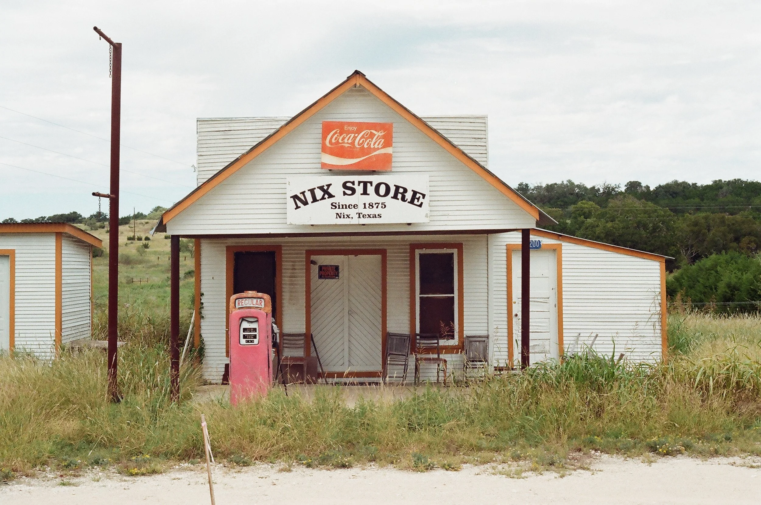 A small white vintage store with a sign that reads 'Nix Store Since 1875 Nix, Texas,' featuring a pink vintage gas pump labeled 'REGULAR' in front. The store has a CocaCola sign on top and is surrounded by tall grass, with a rural landscape in the ba