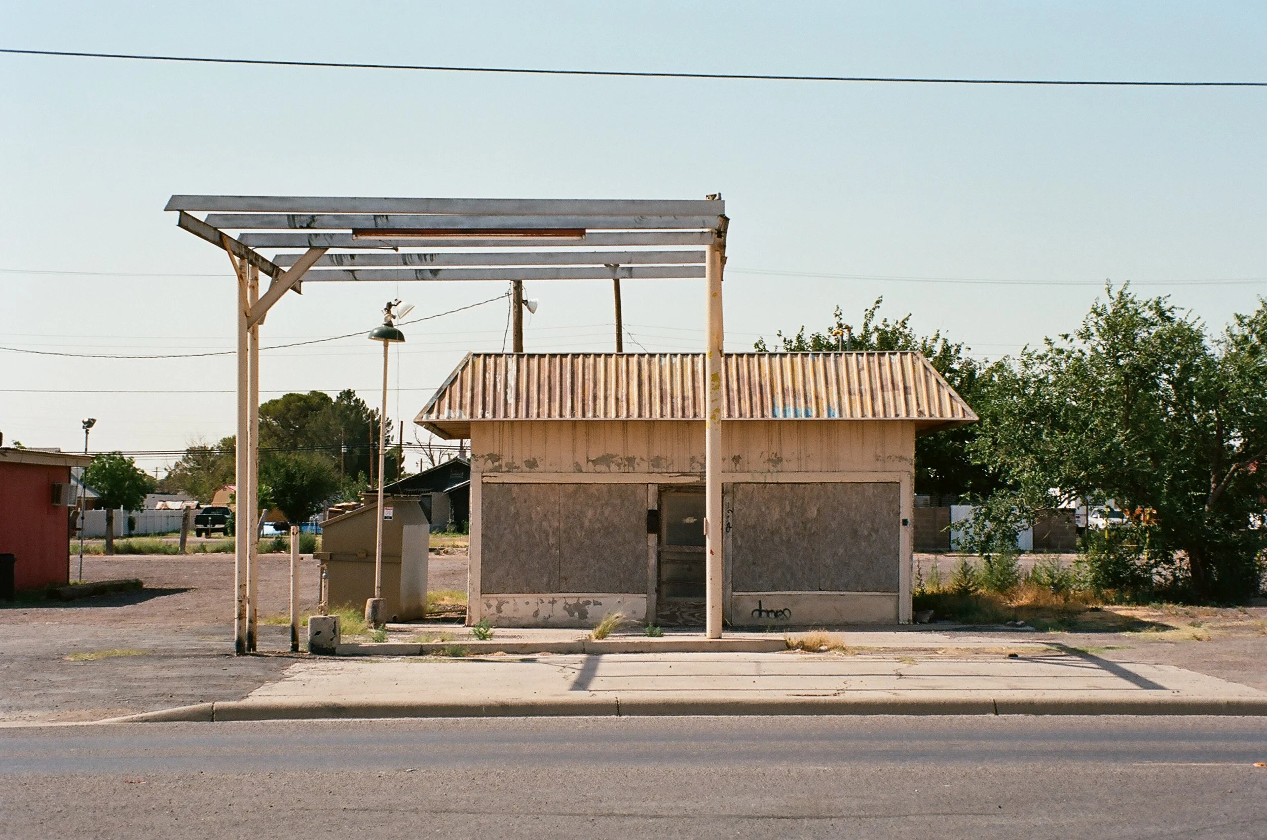 An abandoned, boarded-up small building with a rusty corrugated metal roof, situated on a sidewalk in an empty lot, with a white metal frame structure in front and some trees and other buildings in the background.