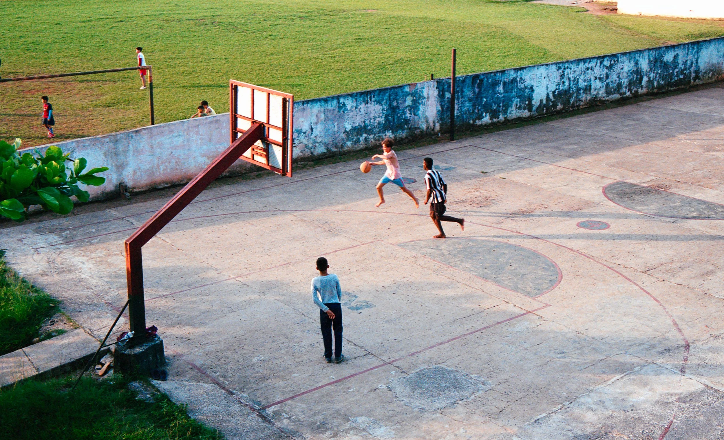 Children playing basketball on an outdoor court while other kids play soccer on a field nearby.