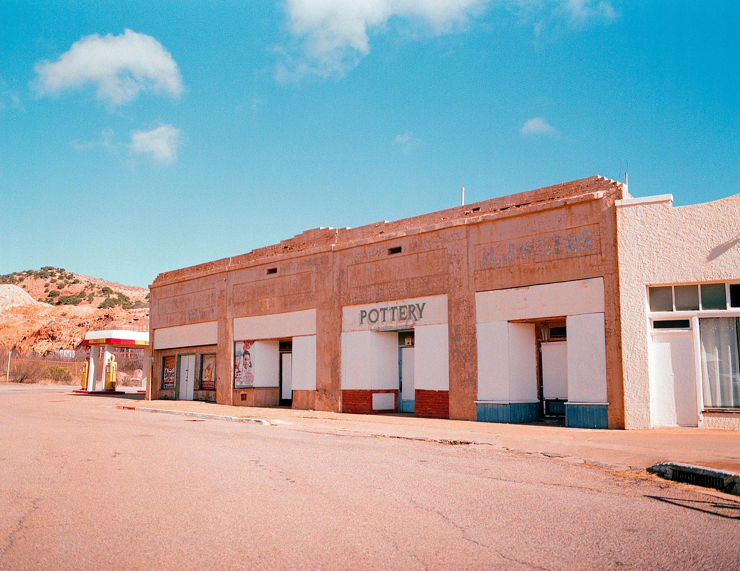 Abandoned building with a sign labeled 'Pottery' on a deserted street, with a gas station visible in the background, under a bright blue sky.