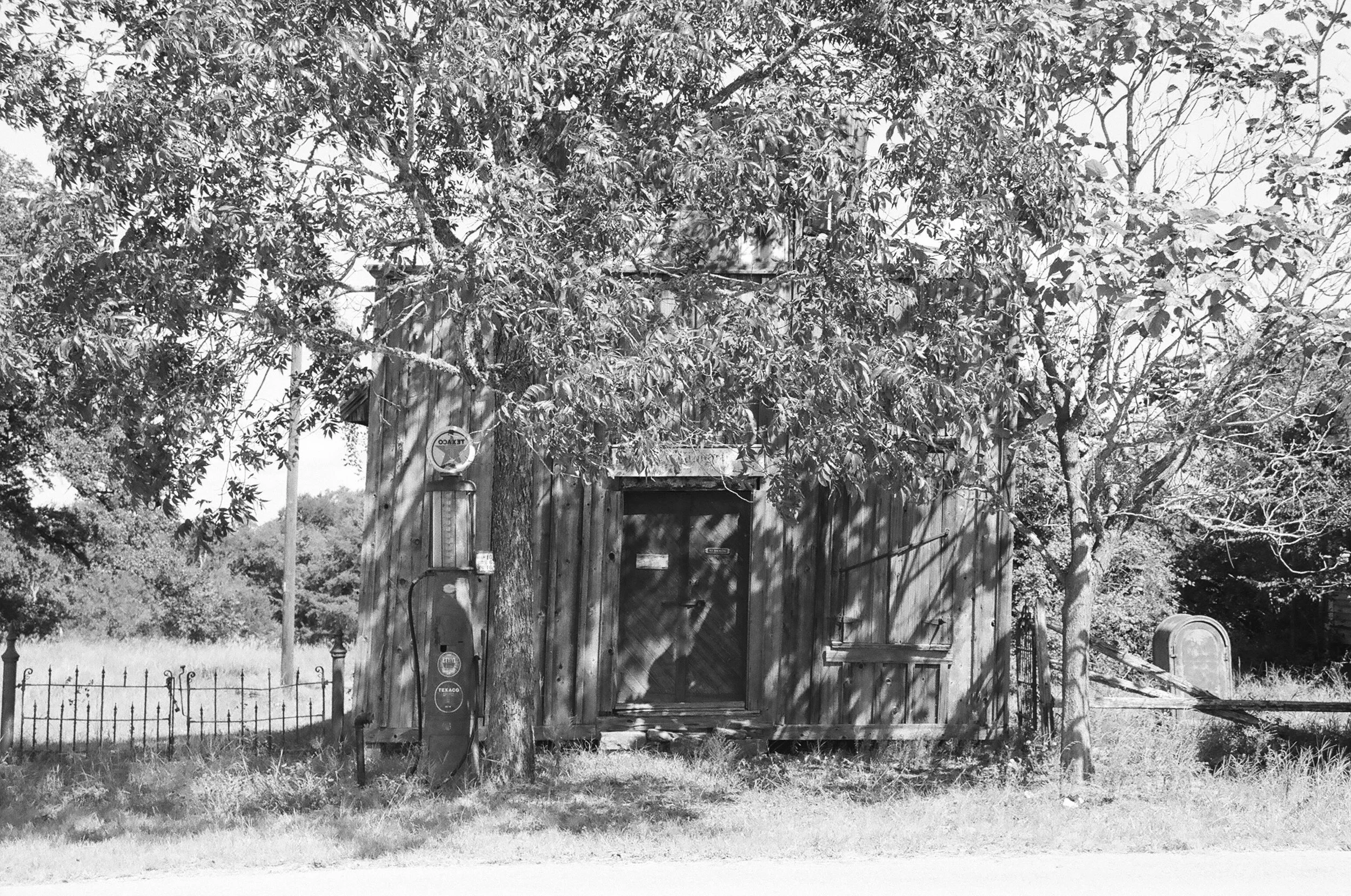 Black and white photo of a rustic wooden shed with trees and overgrown grass surrounding it, an old gas pump is visible on the left side.