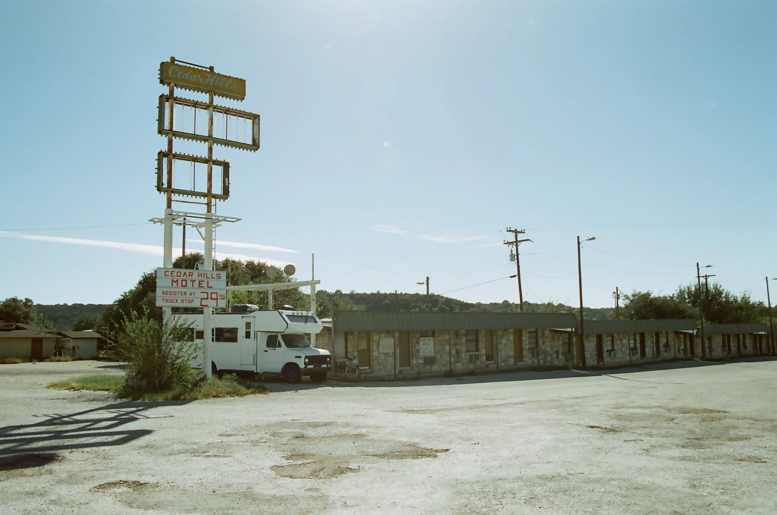 An empty parking lot with a sign for Cedar Hills Motel and an RV parked nearby, under a clear blue sky.