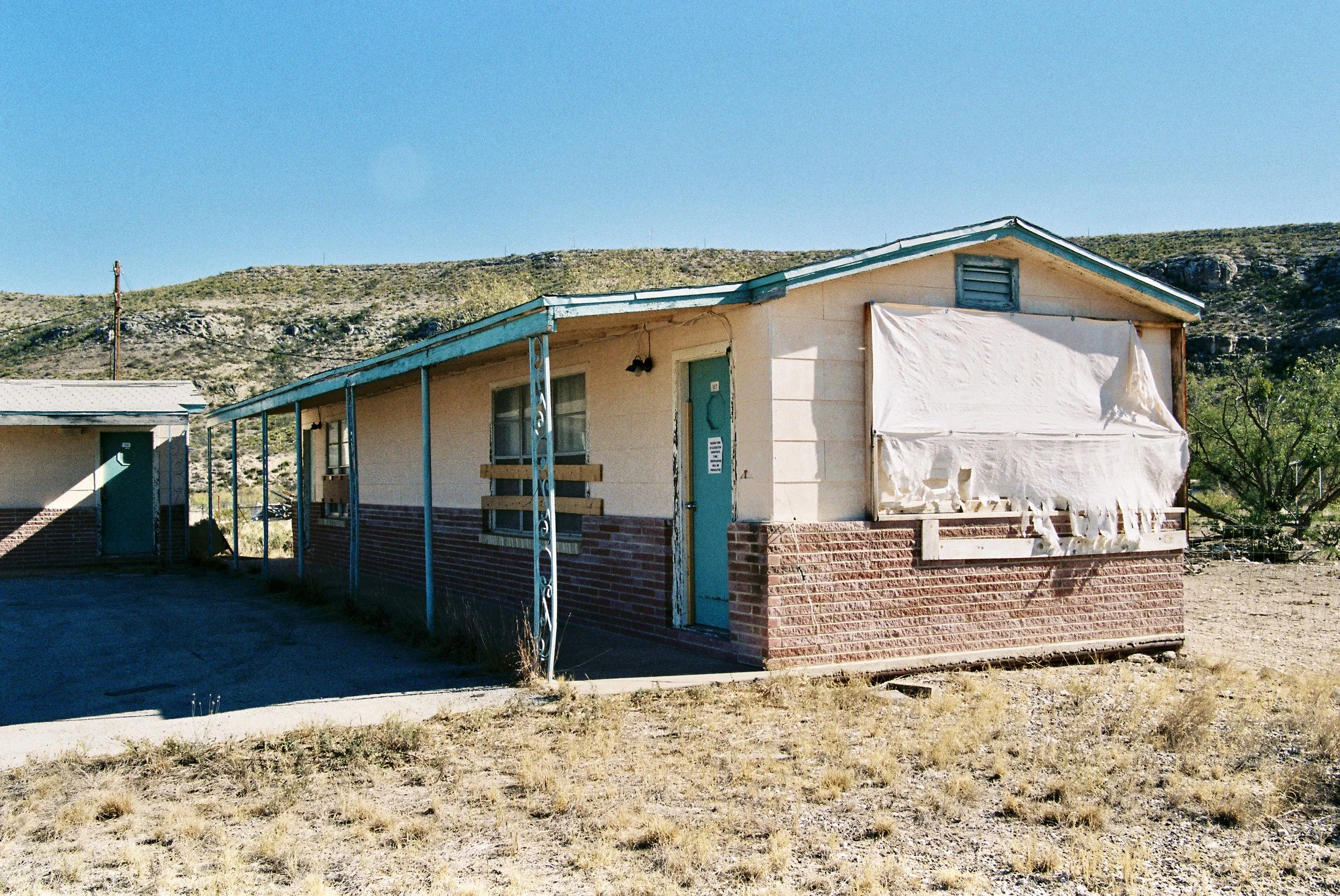 A small, weathered house in a dry desert landscape under a clear blue sky. The house has a blue door, brick base, and a makeshift cover over the window on the right side.