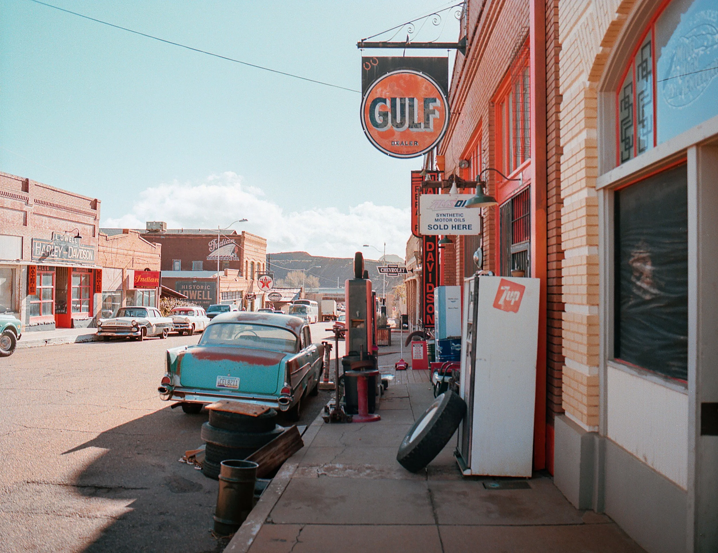 A vintage small-town street scene with old cars, gas pumps, and nostalgic storefronts, featuring signs for Gulf and Harley-Davidson.