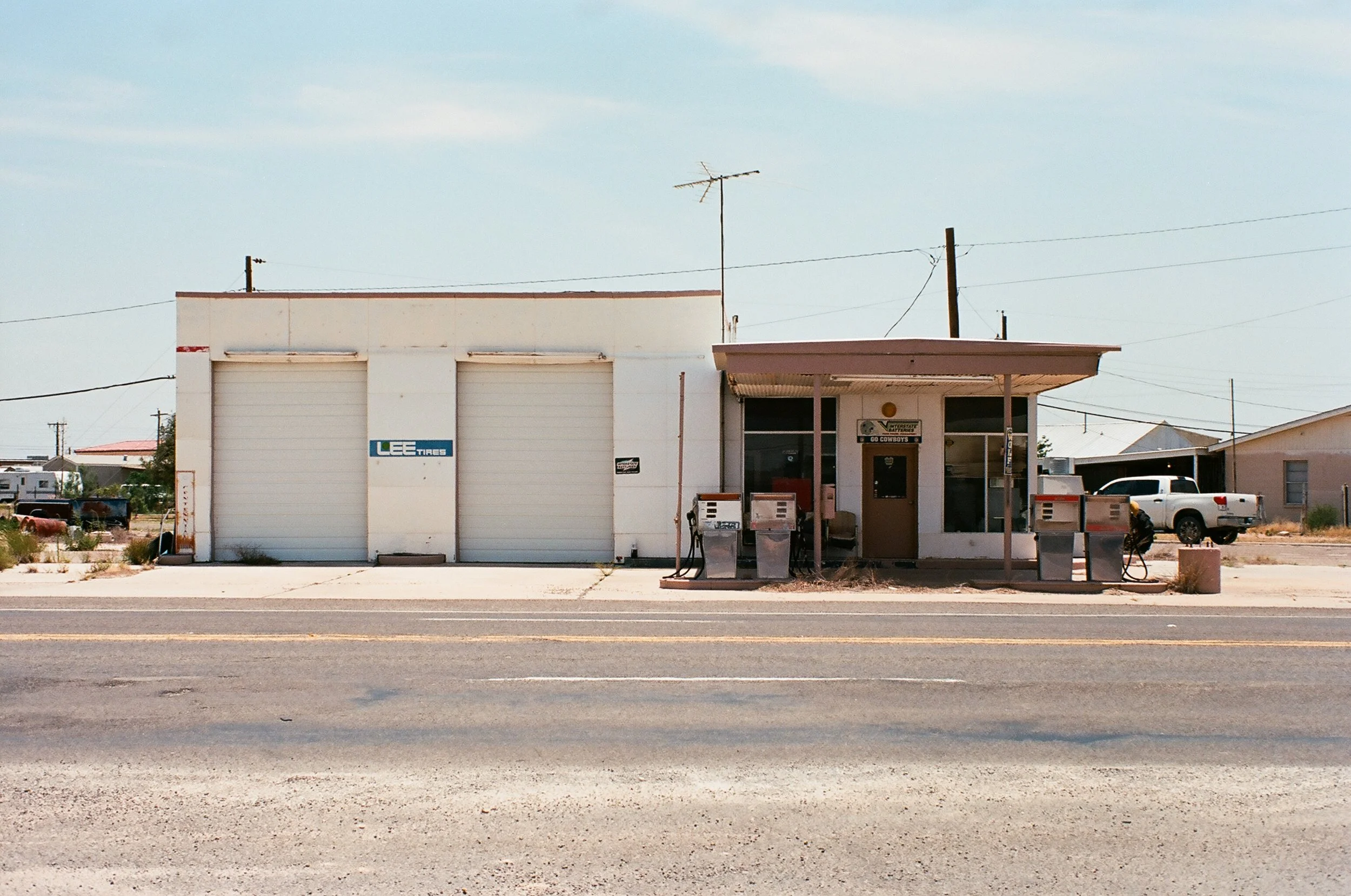Old, weathered gas station with two gas pumps outside, a small office with a door and windows, and an attached garage with a "Lee Tires" sign, located on a deserted street.