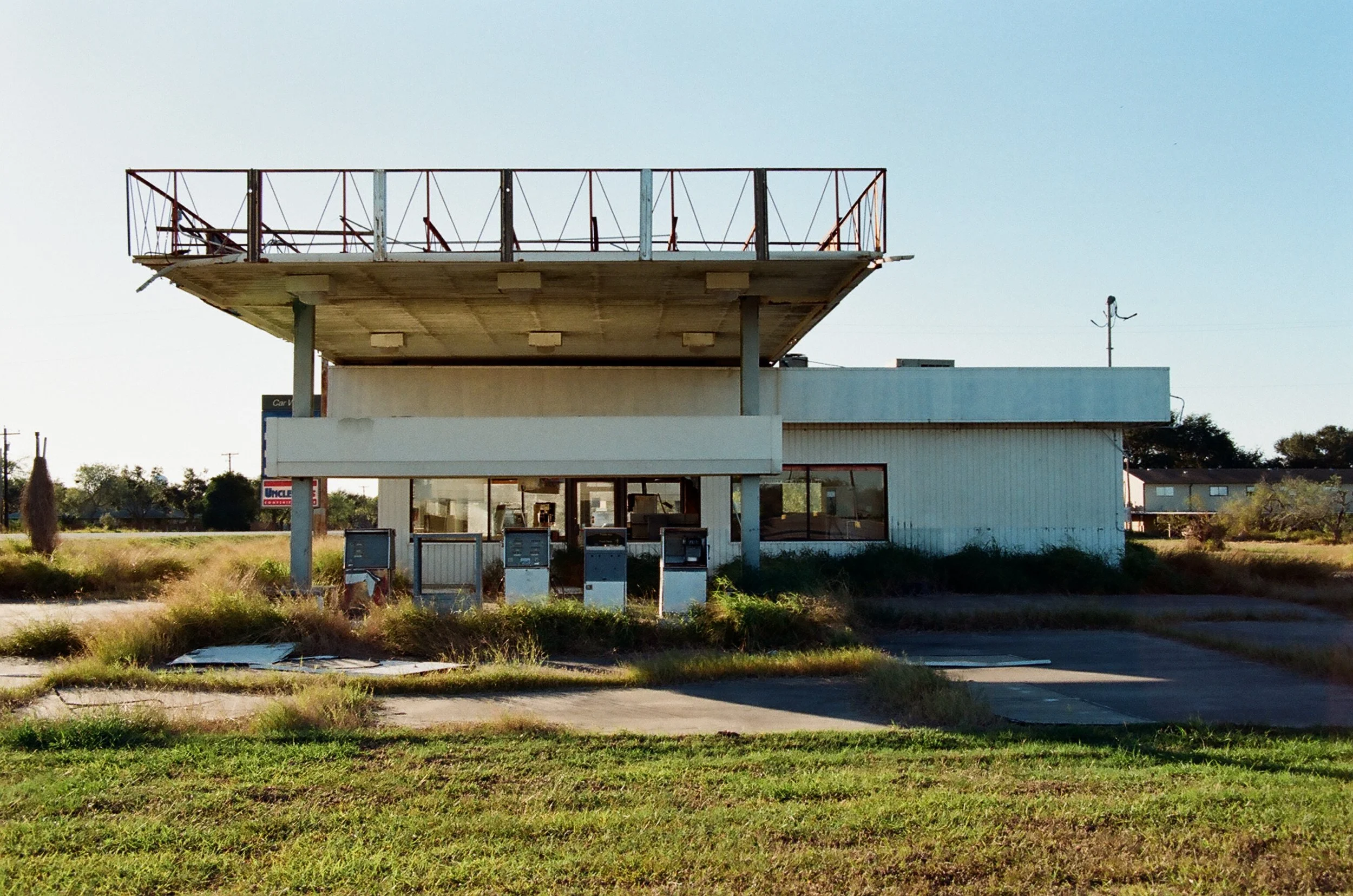 Abandoned building that looks like an old fast-food restaurant with a large empty balcony or roof area on top, surrounded by overgrown grass and weeds.