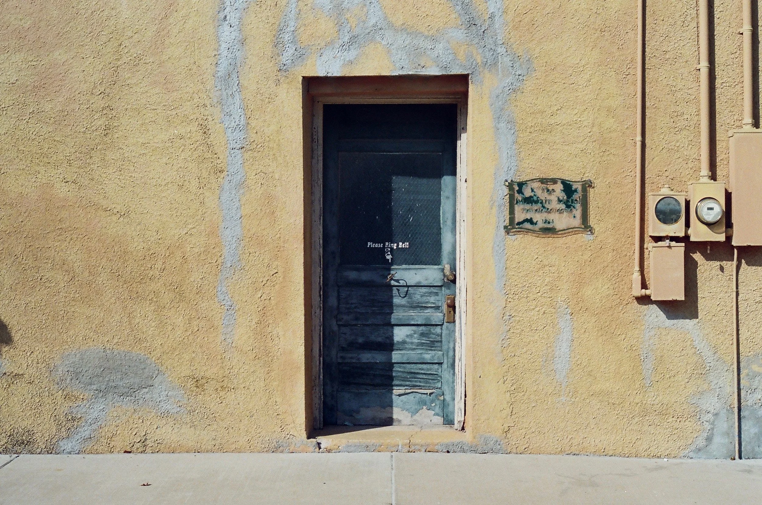 Old blue wooden door on a yellow textured wall with a small sign that reads 'Please Ring Bell' and old electrical meters on the side.