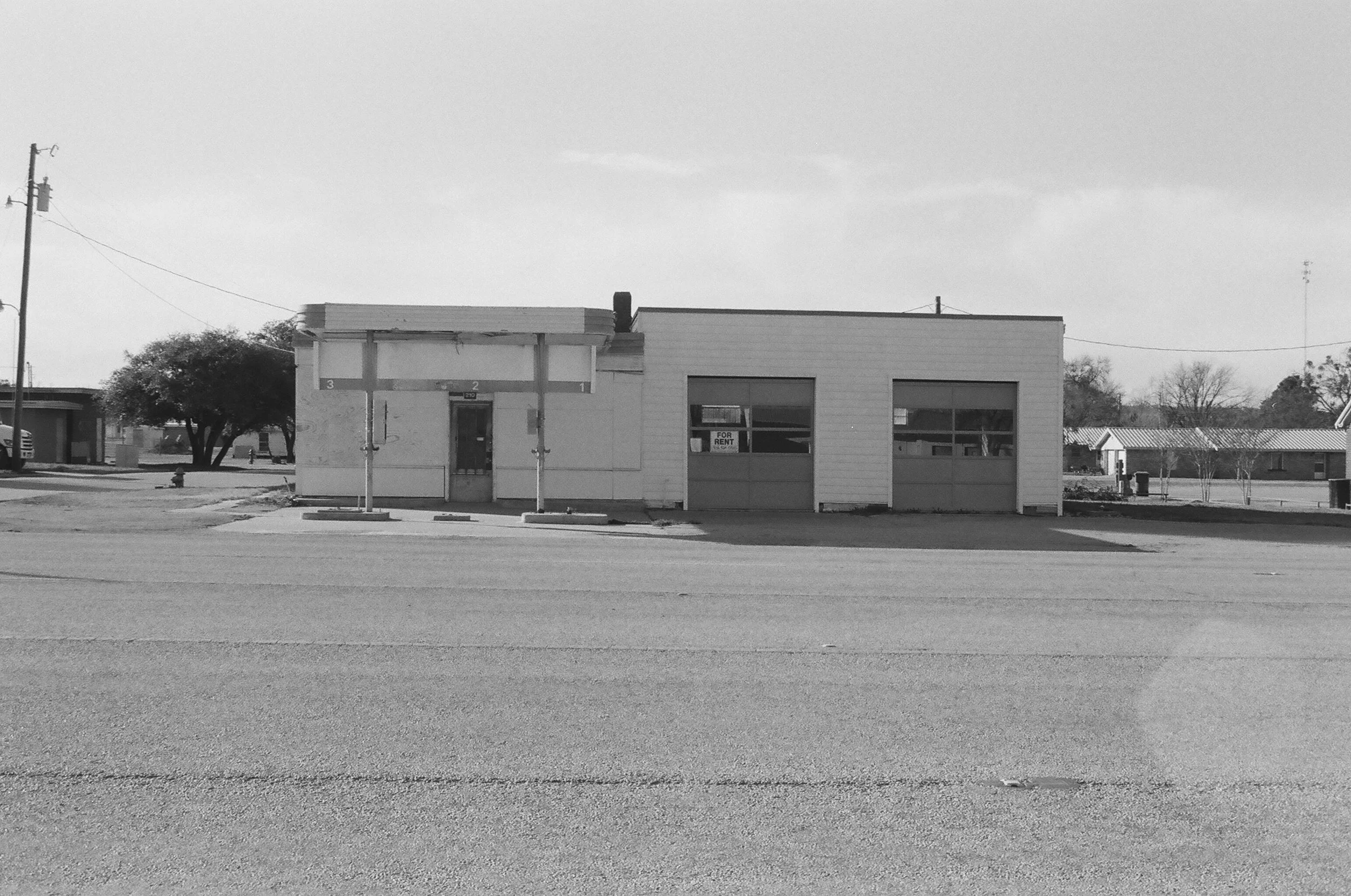 Empty commercial building with a sign in the window that reads 'For Rent,' and a parking lot in front.