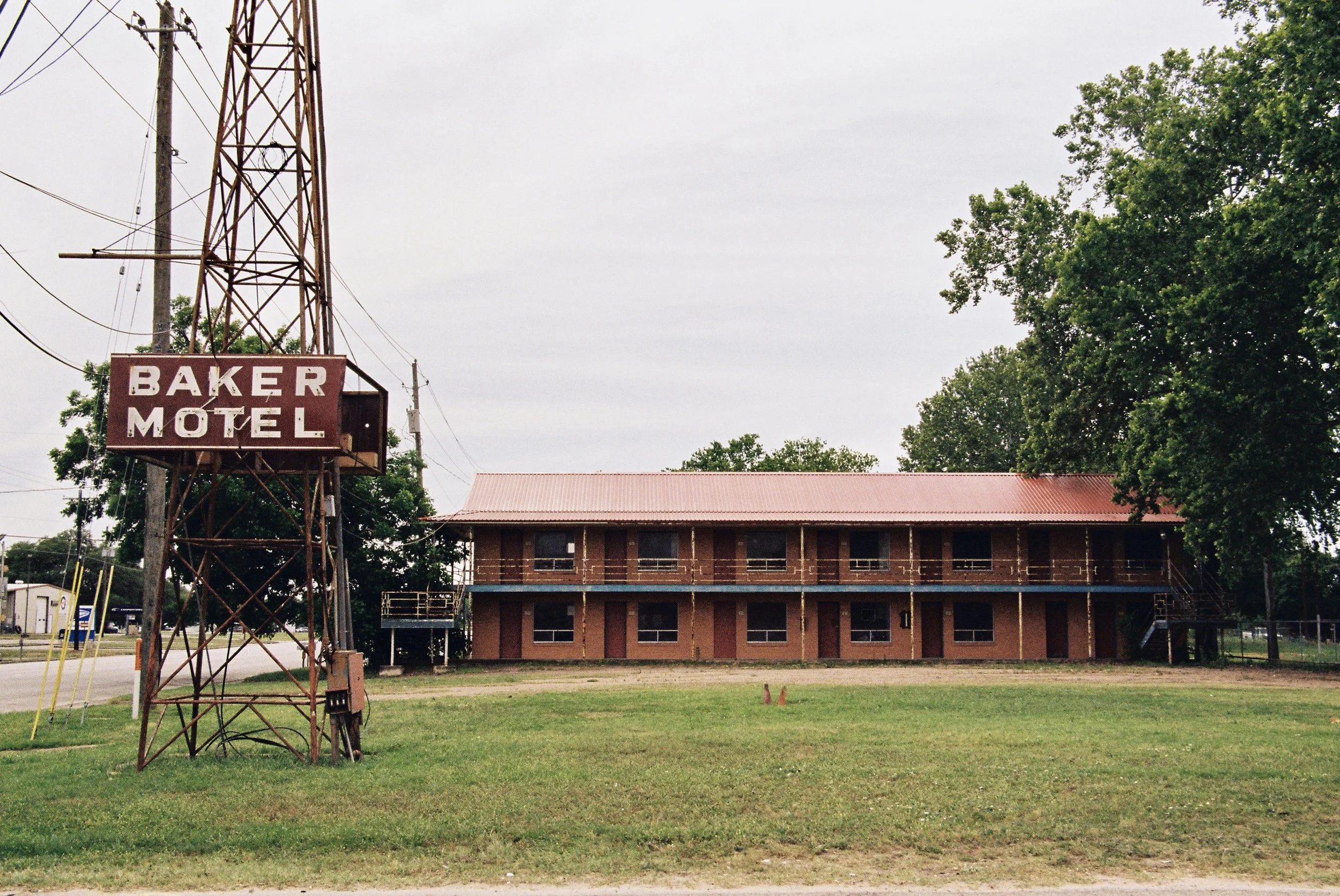 An old two-story motel building with a red metal roof and a large sign that reads 'Baker Motel' on a tall, rusted metal tower. The motel has a brick exterior and a balcony on the second floor, with stairs on both sides. There is a grassy field in fro