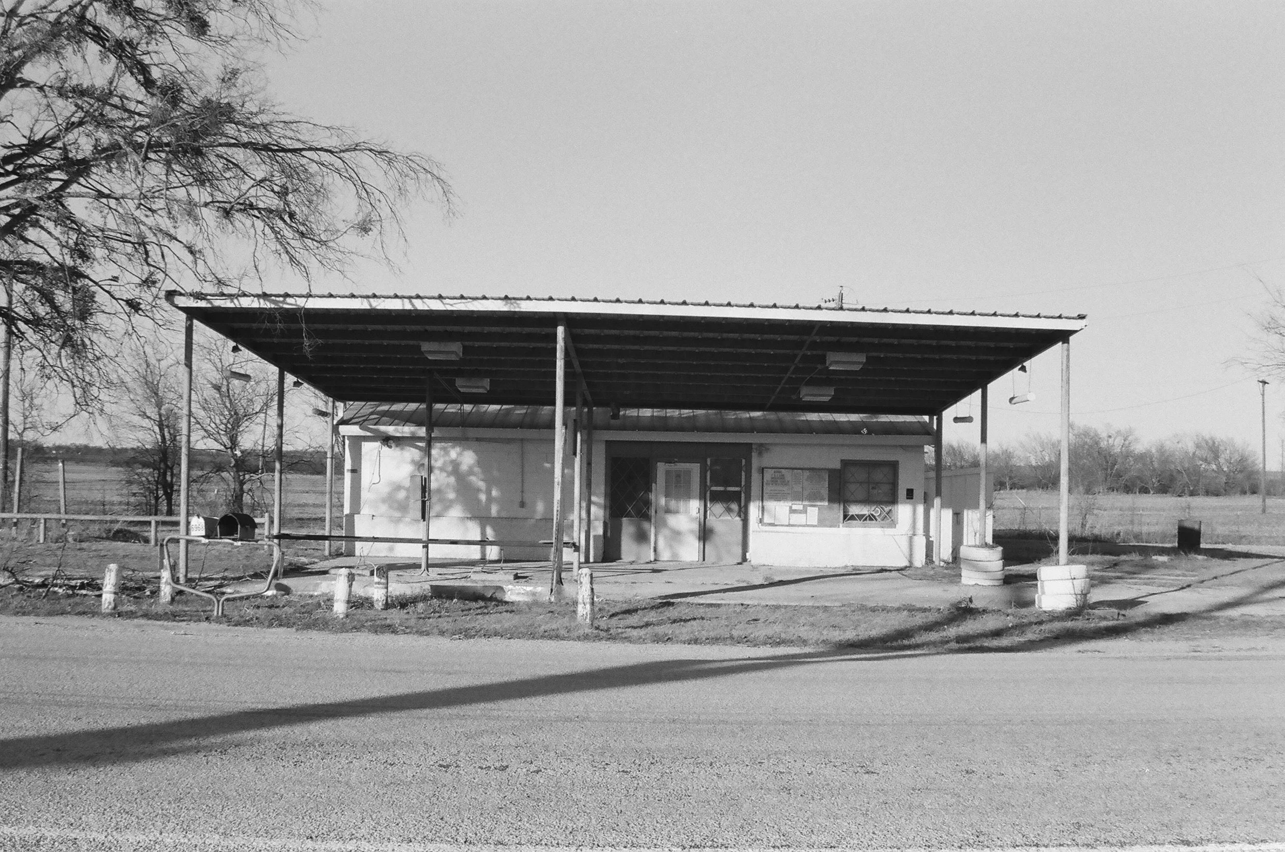 A small, old, building with a metal roof and a covered porch, appears abandoned with no people around. The scene is in black and white and depicts a rural setting with bare trees and open fields in the background.