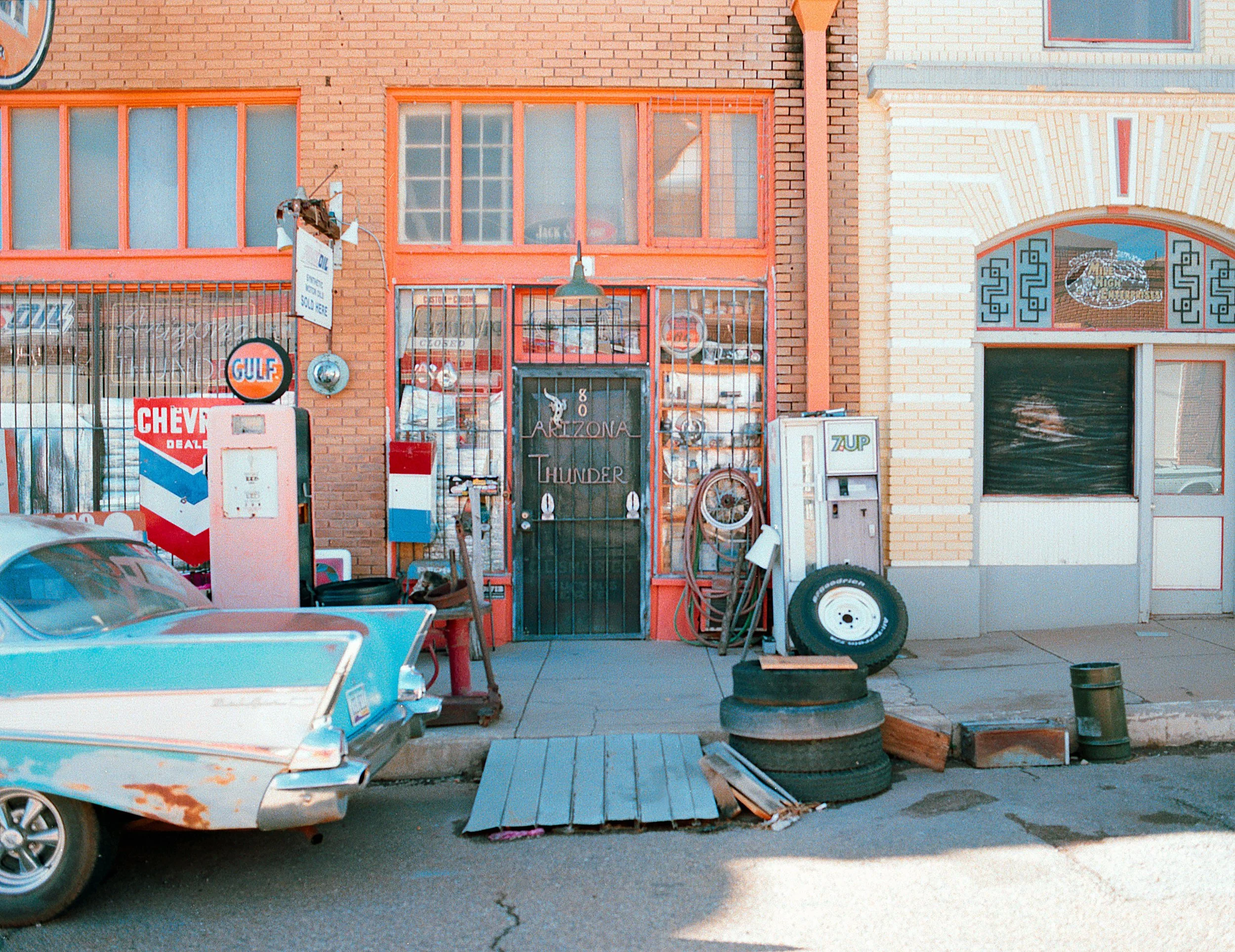 Old vintage car parked in front of a small auto repair shop with gas pumps, tires, and tools outside, in a colorful building with signs and windows.