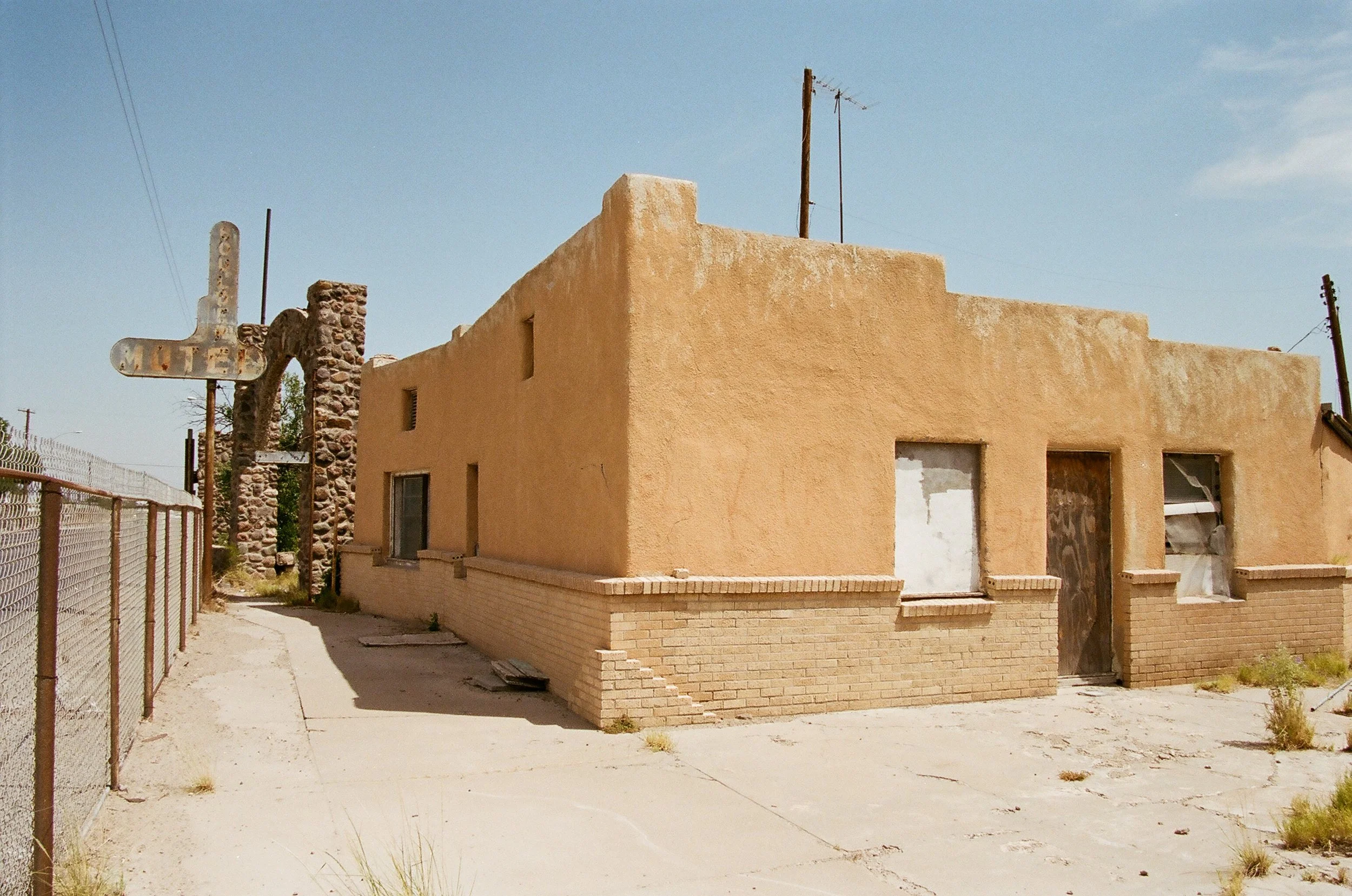 An abandoned building with a tan stucco exterior and boarded-up windows, located on a dirt lot with sparse vegetation, in a rural area under a clear sky.