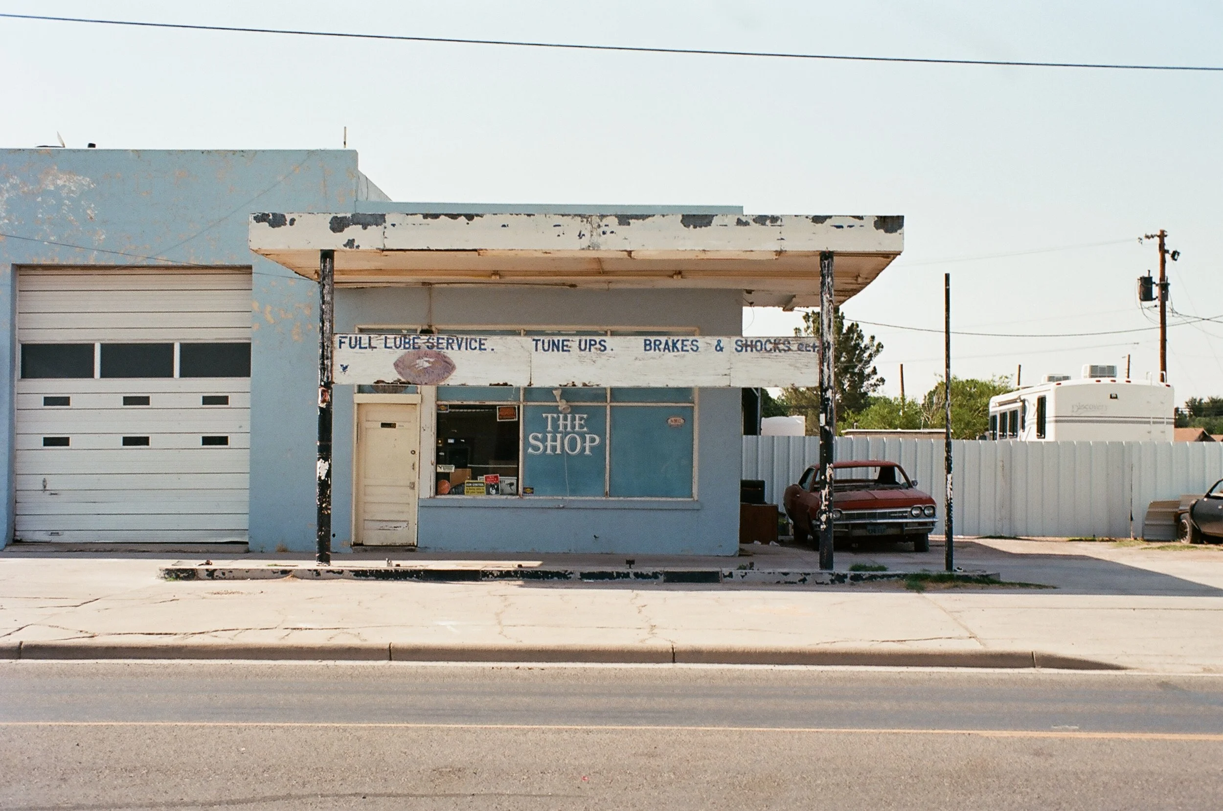 An old, weathered auto repair shop named 'The Shop' with a faded blue exterior and peeling paint. There is a red vintage car parked under the front shelter. The storefront has a sign offering full lube service, tune-ups, brakes, and shocks. A white w