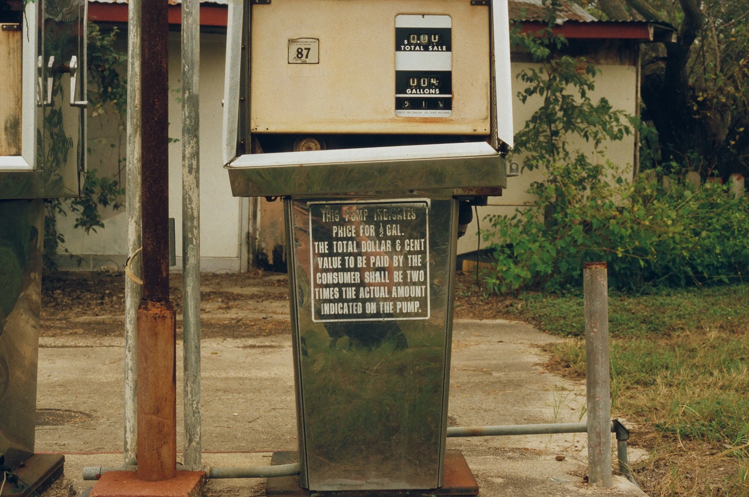 Vintage gas pump with handwritten instructions, standing on a concrete sidewalk with grass and trees in the background.