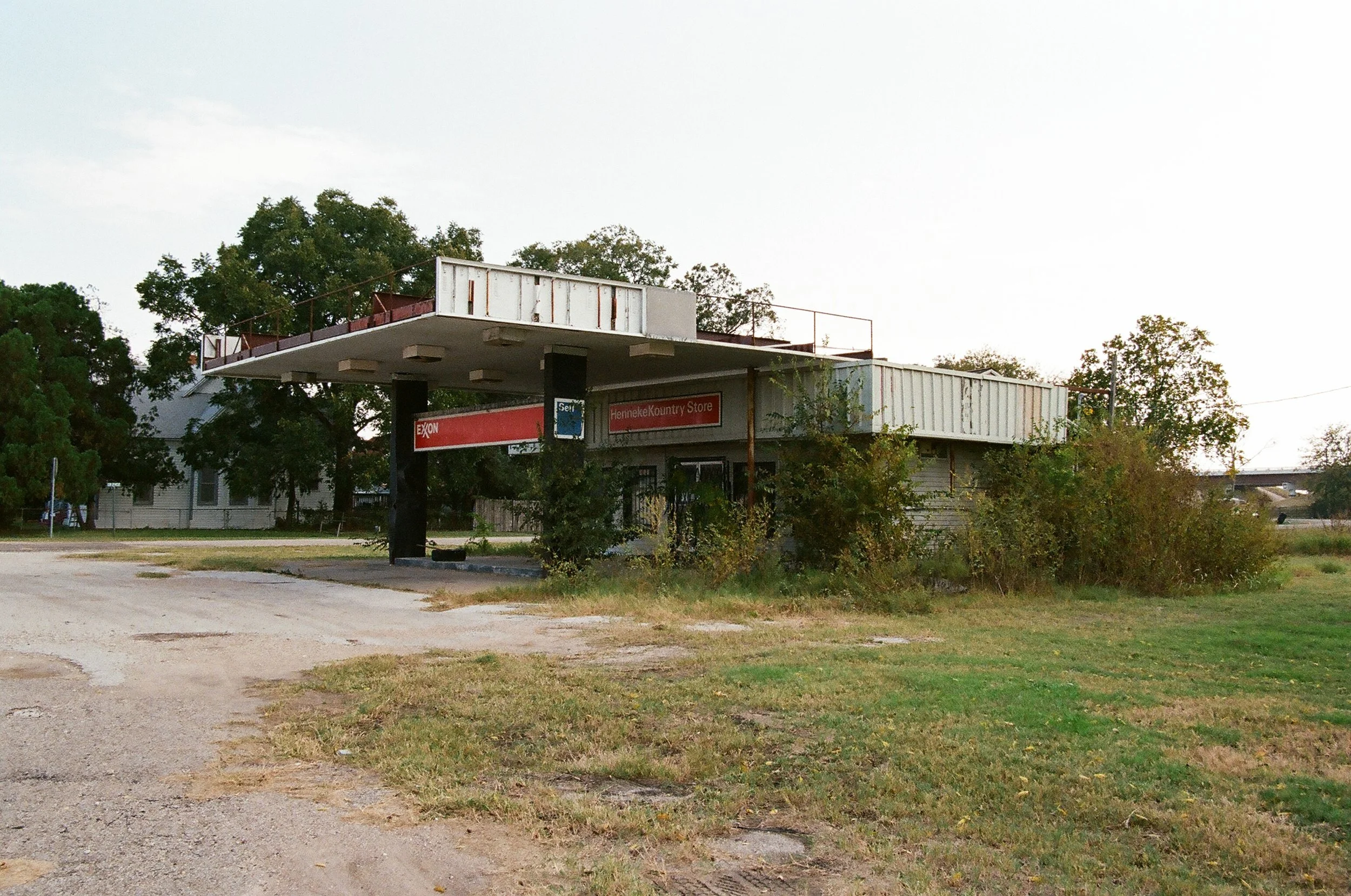 An abandoned and dilapidated Shell gas station with overgrown shrubbery and worn signage, situated on a grassy area with a dirt patch in the foreground.