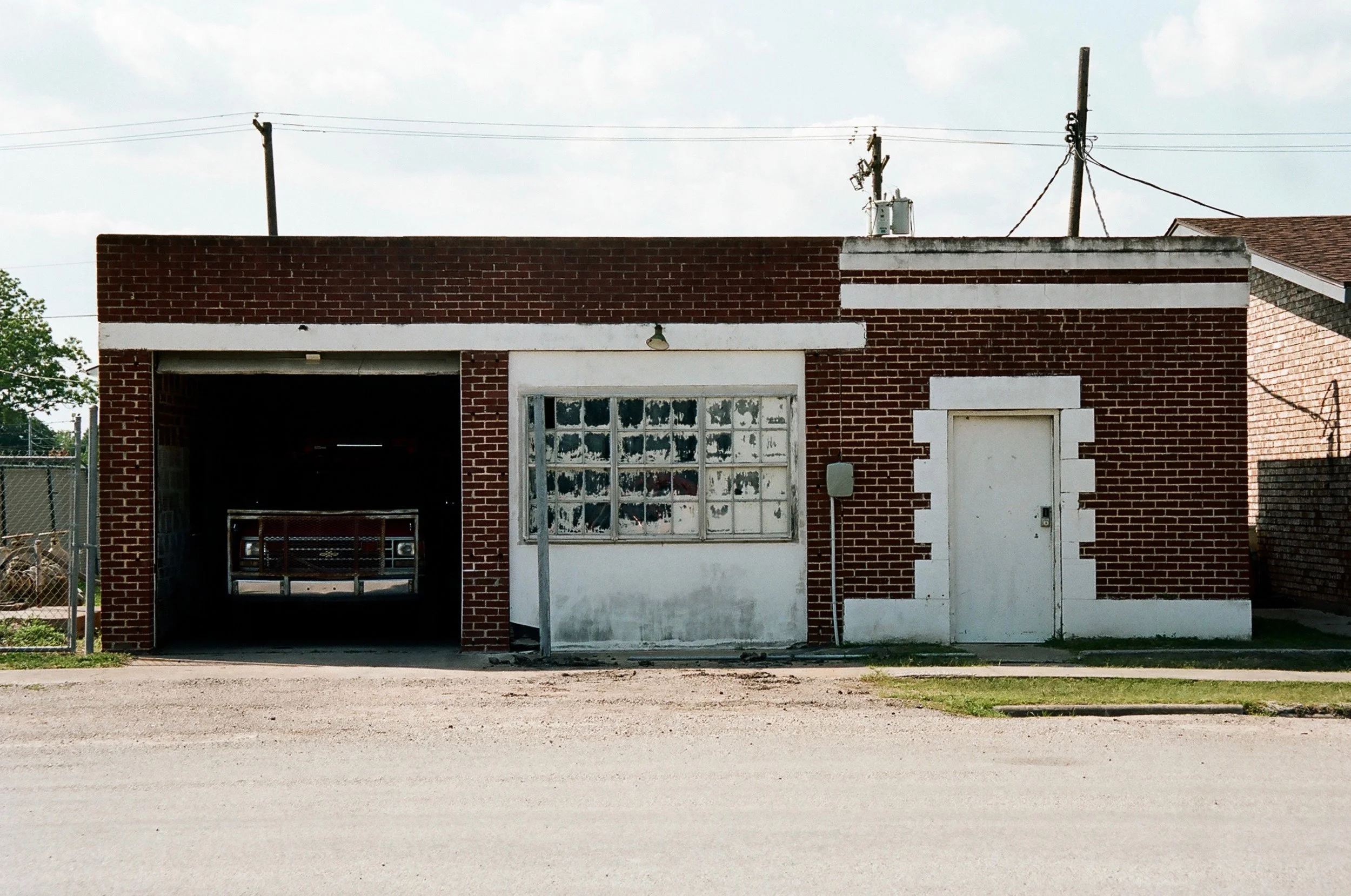 A small brick building with a garage on the left, a window in the middle, and a door on the right. The garage contains a vehicle with a front grille visible. The window appears to be damaged or aged, with peeling paint. The building has utility wires