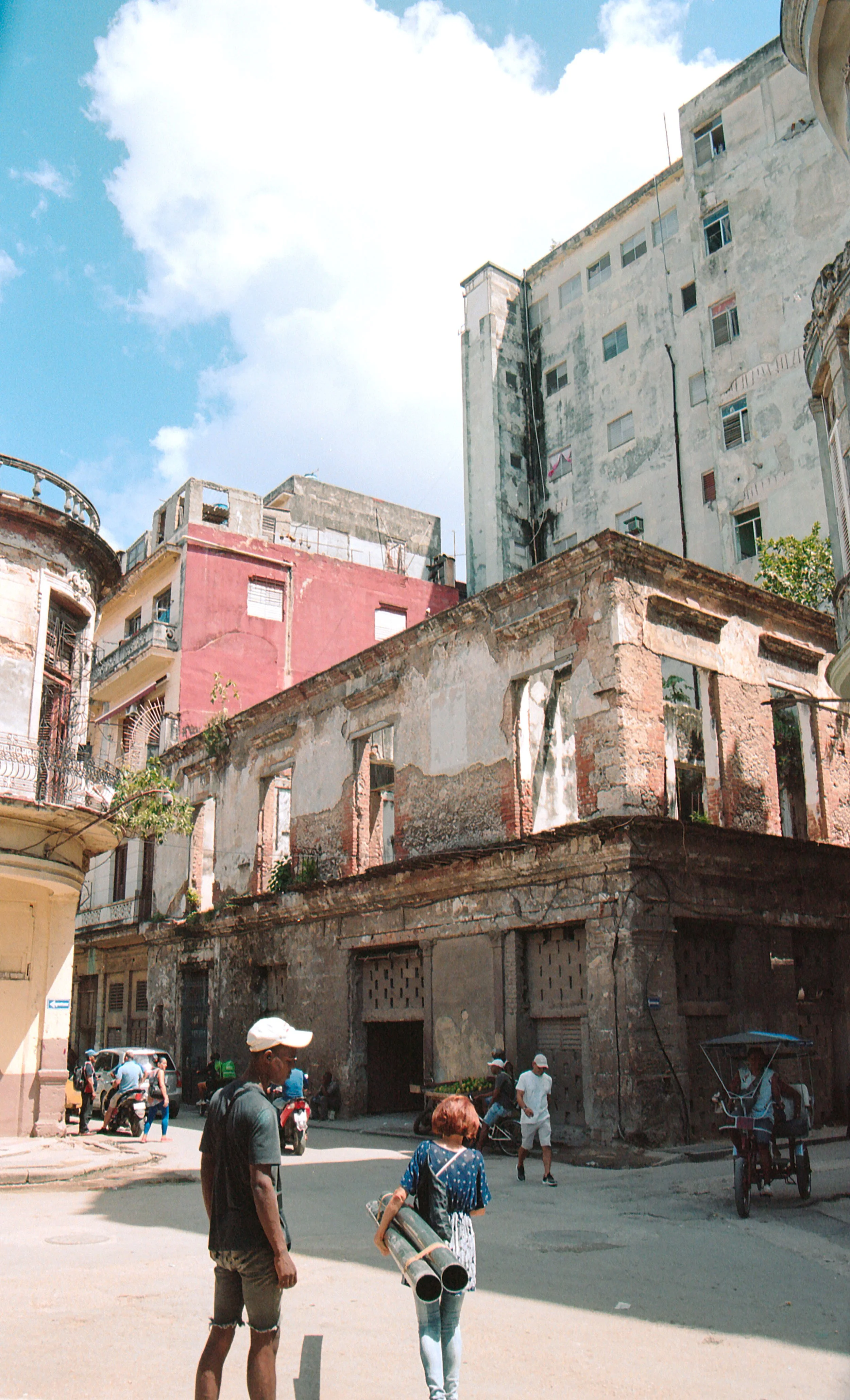 Group of people walking and riding bikes on a street with old, weathered buildings in the background.