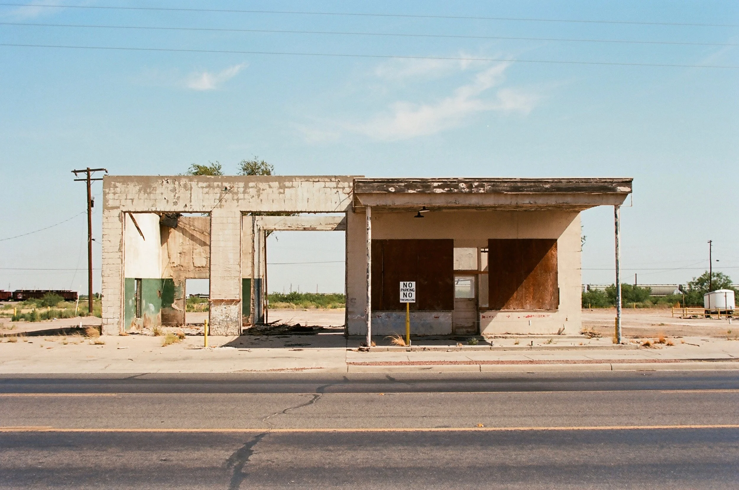 An abandoned, rundown building with boarded-up windows and peeling paint, located in a desert-like area with sparse vegetation. There is a 'No Parking' sign in front of the building and electrical wires overhead.