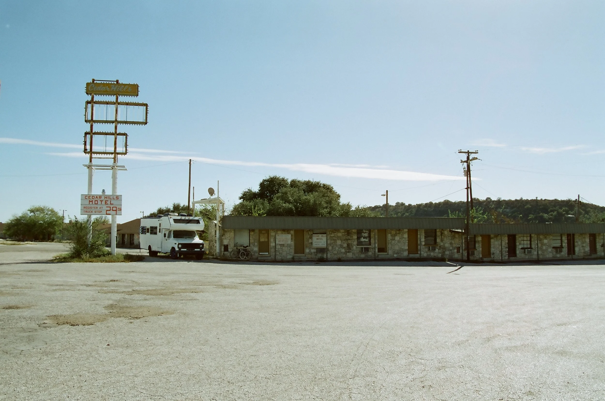 Empty parking lot in front of a small stone motel building with a sign that reads 'Cedar Hills Motel', a bus parked nearby, and utility poles along the road in a rural area on a clear, sunny day.