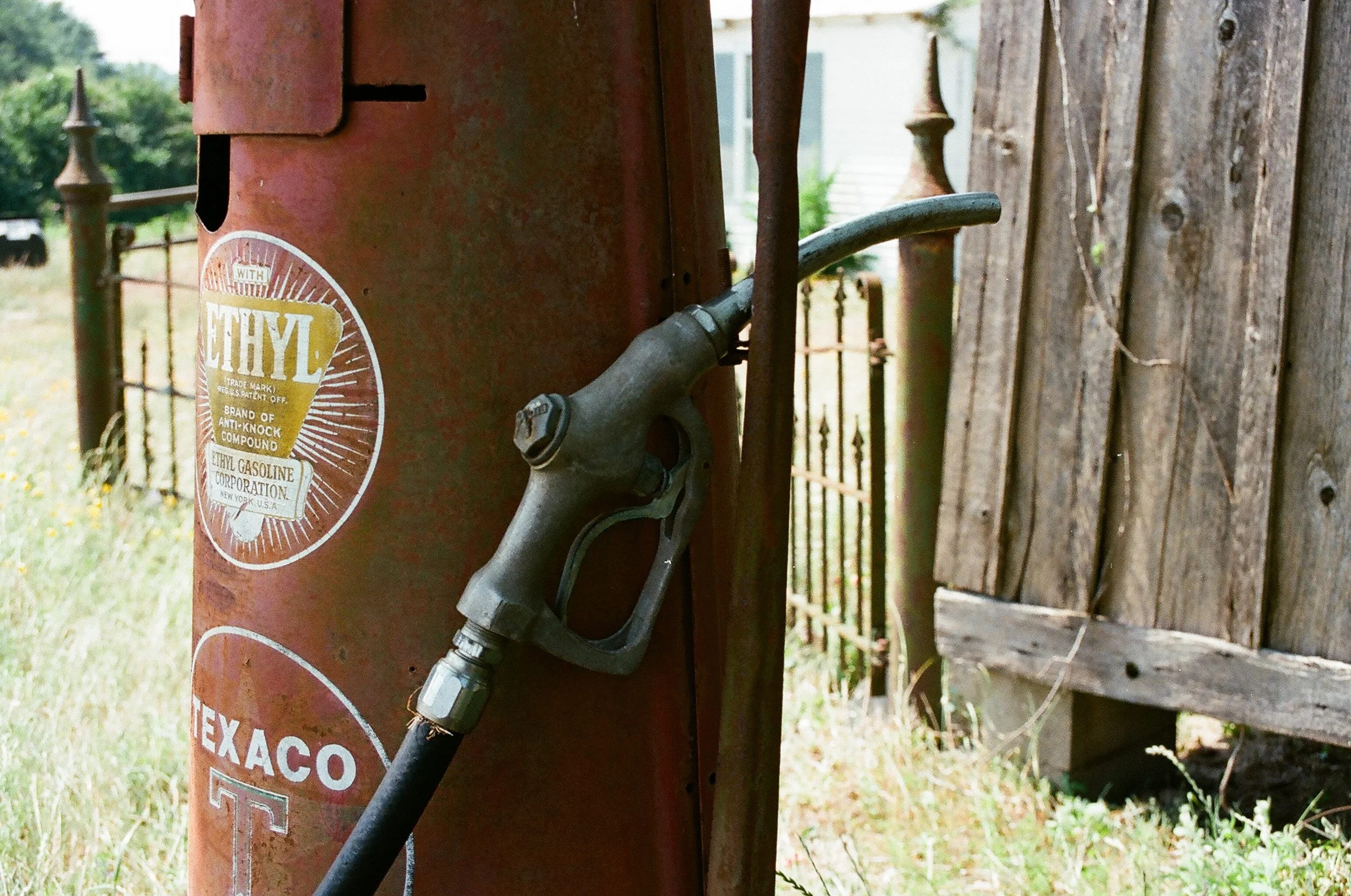 An old gasoline pump with a vintage Texaco and Ethyl sticker, attached to a rusty metal stand next to a weathered wooden fence.