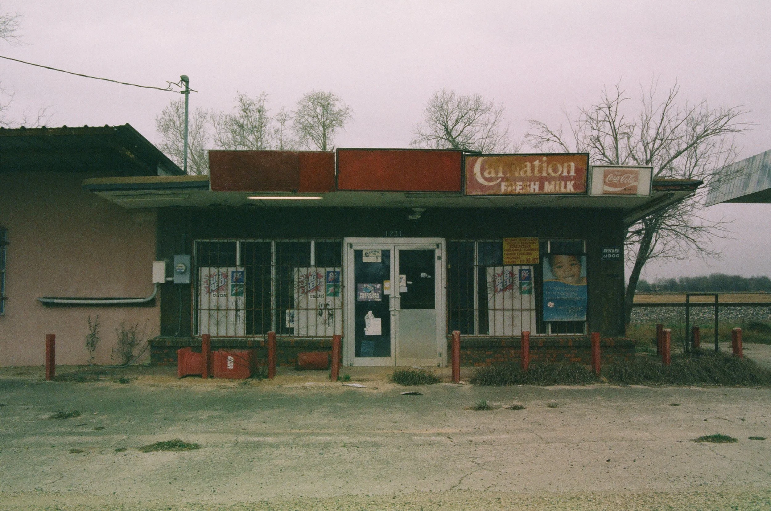 Small, old convenience store with red and black signage, metal security gates over windows, surrounded by a gravel lot, with a tree and open field in the background.