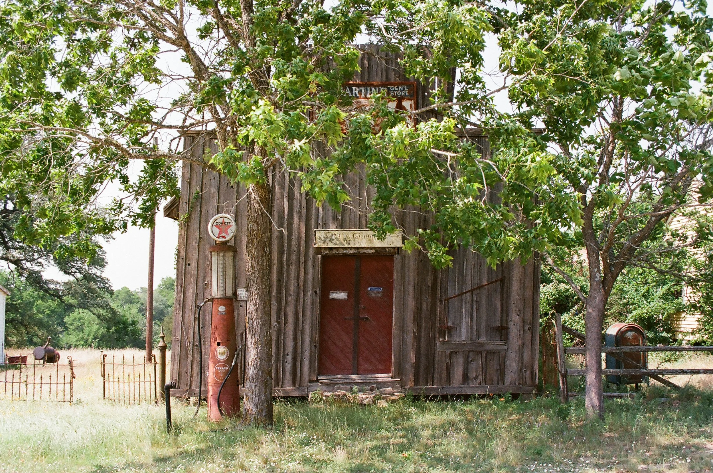 An old wooden building with a red door, surrounded by trees and grass, with a vintage gas pump in front.