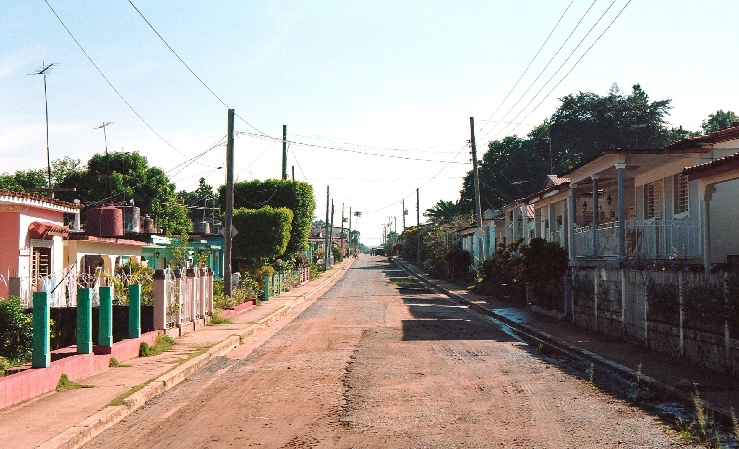 A quiet residential street with colorful houses, a dirt road, and utility poles under a clear sky.