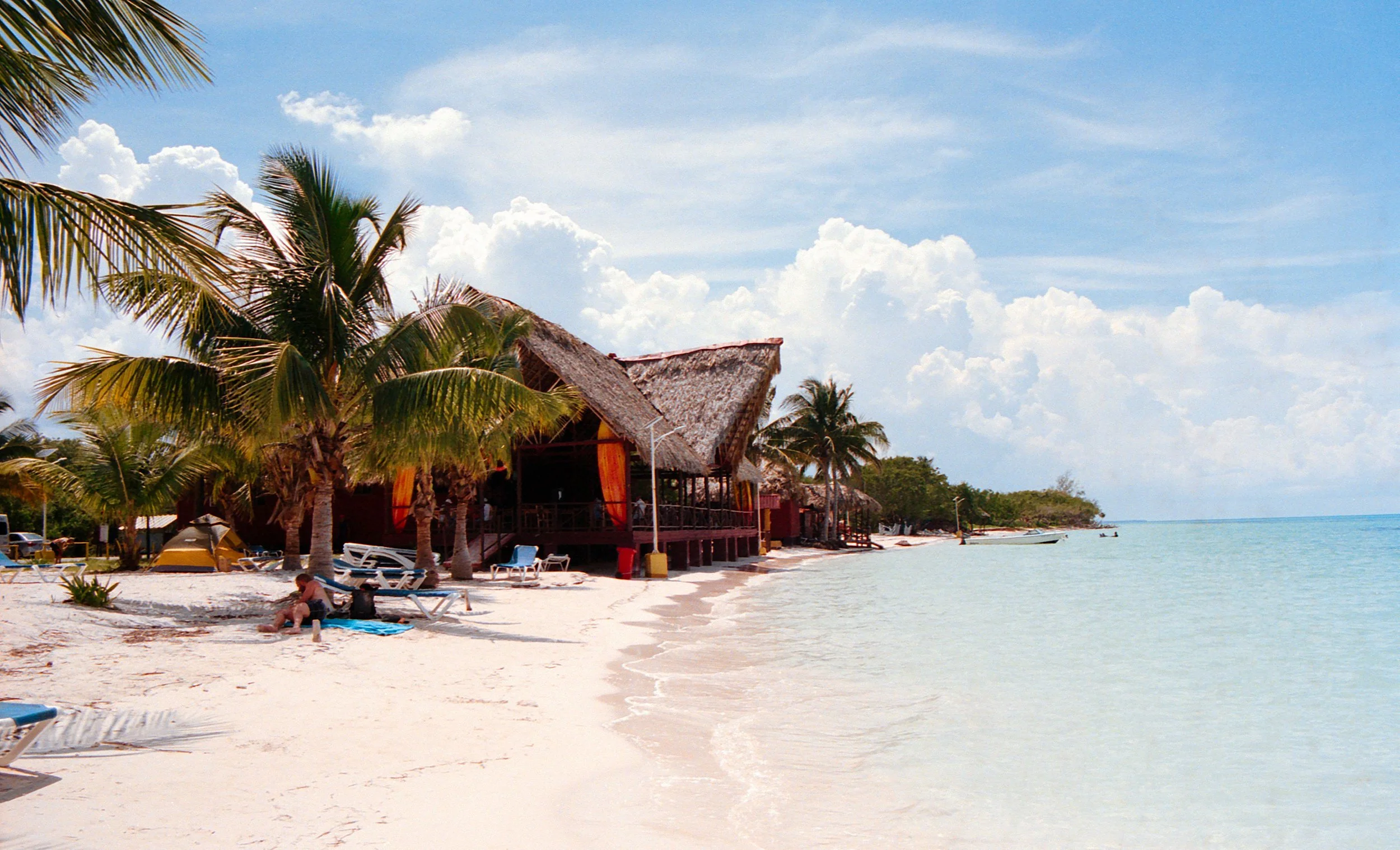 Beach scene with palm trees, thatched-roof huts, lounge chairs, and clear turquoise water under a partly cloudy sky.