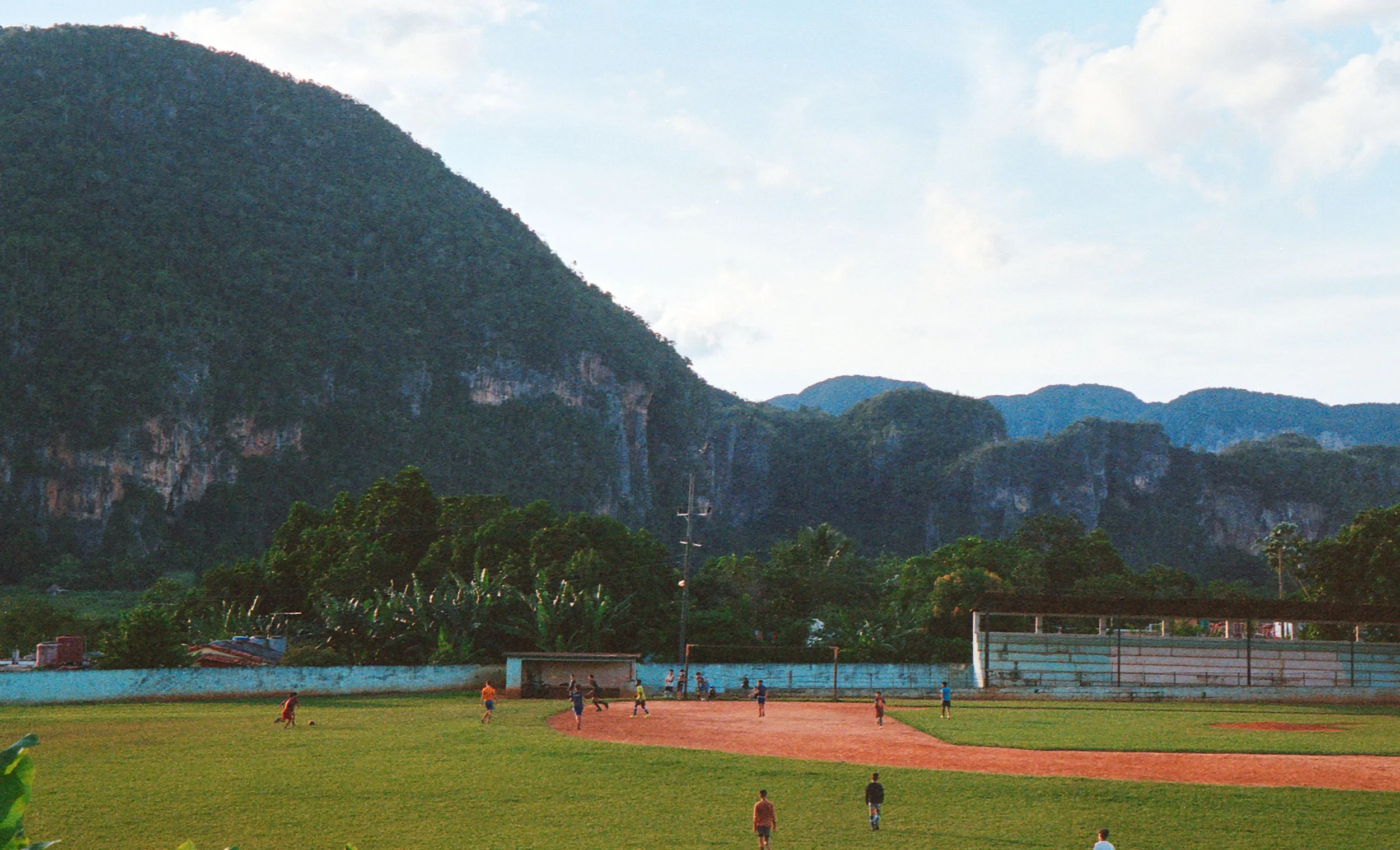 Children playing football on a field with a mountain backdrop and trees.