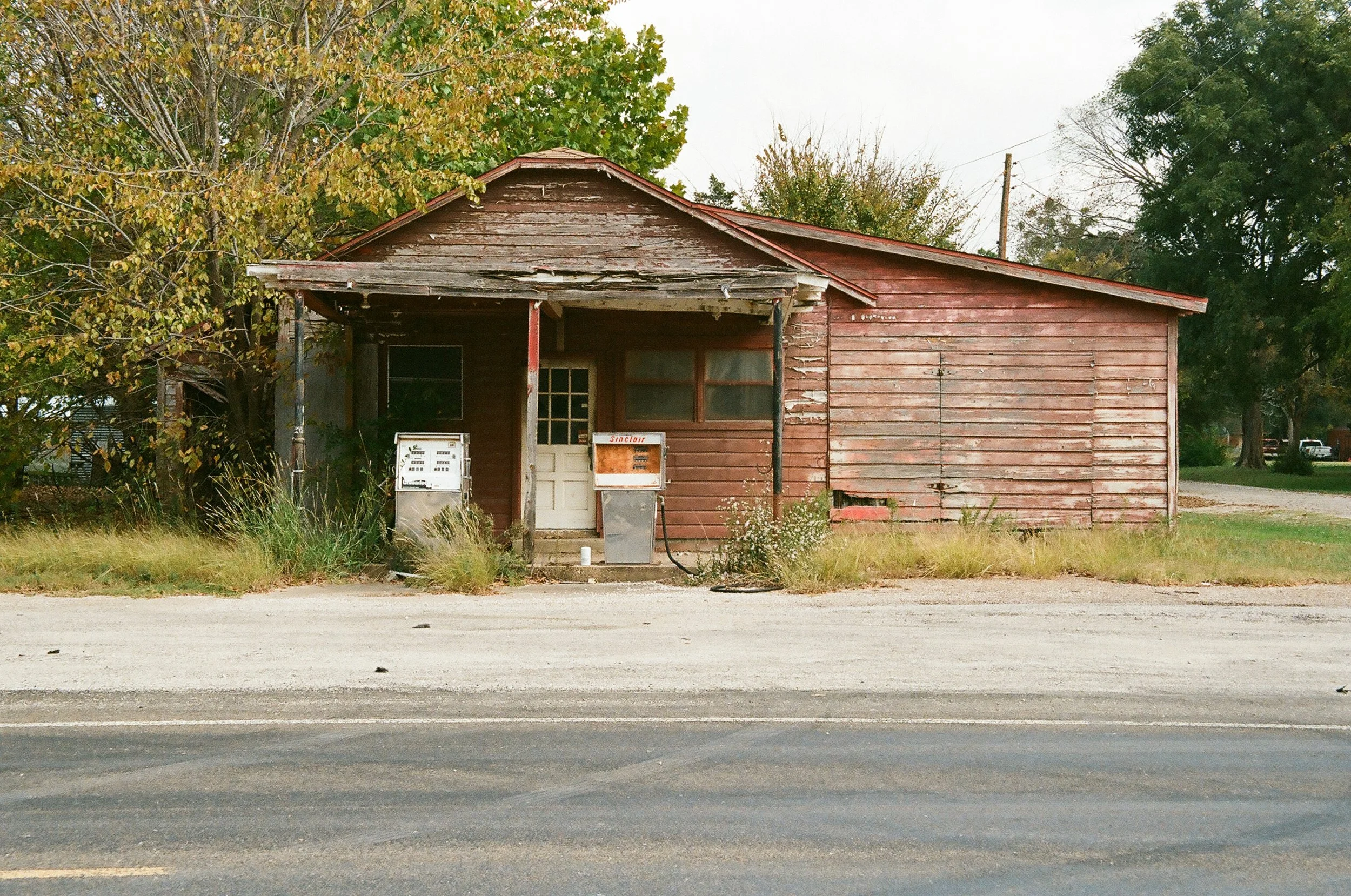 Old, weathered wooden building with peeling red paint, overgrown grass in front, and two vending machines outside, with trees and power lines in the background.