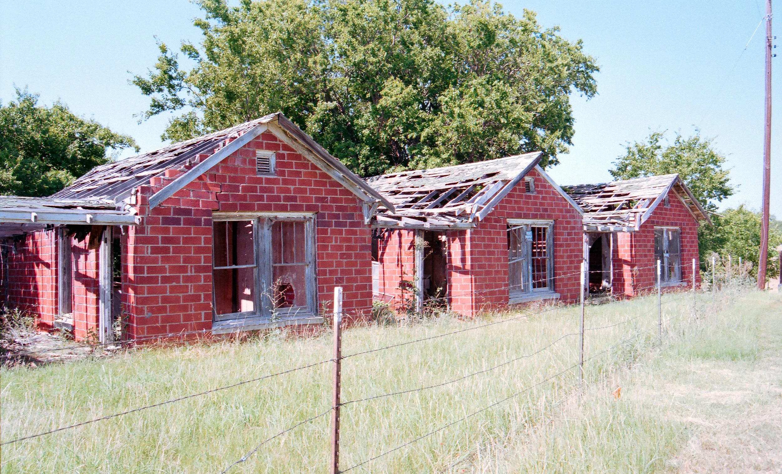 Old, abandoned brick houses with broken windows and sagging roofs behind a wire fence, with green grass and trees in the background.