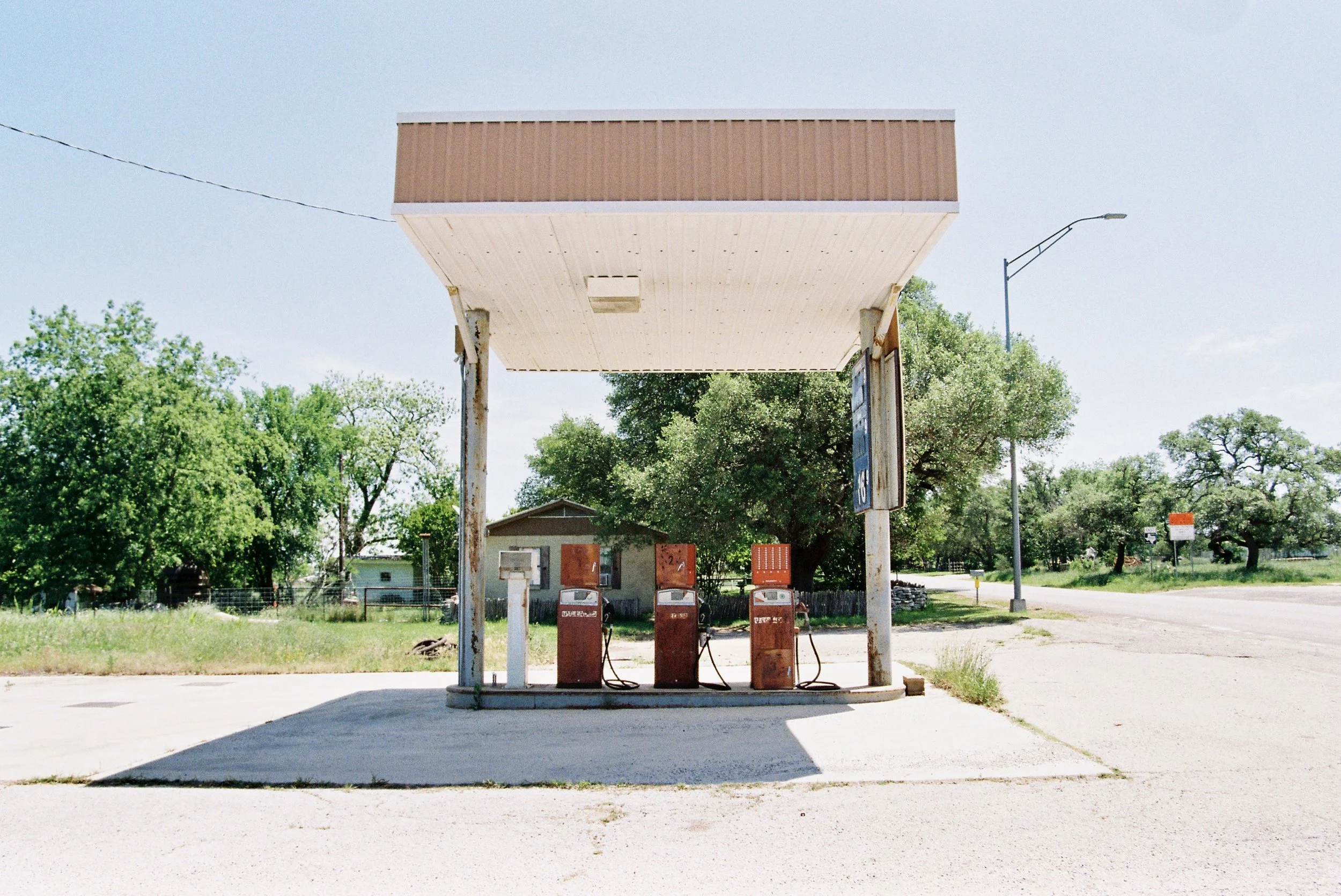 Old, rusty gas pumps at a small, abandoned gas station with a partial roof and a concrete parking area, surrounded by trees and a rural setting.